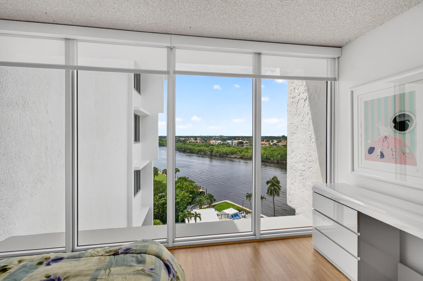 Bedroom with floor-to-ceiling windows overlooking a waterway and palm-lined shore.