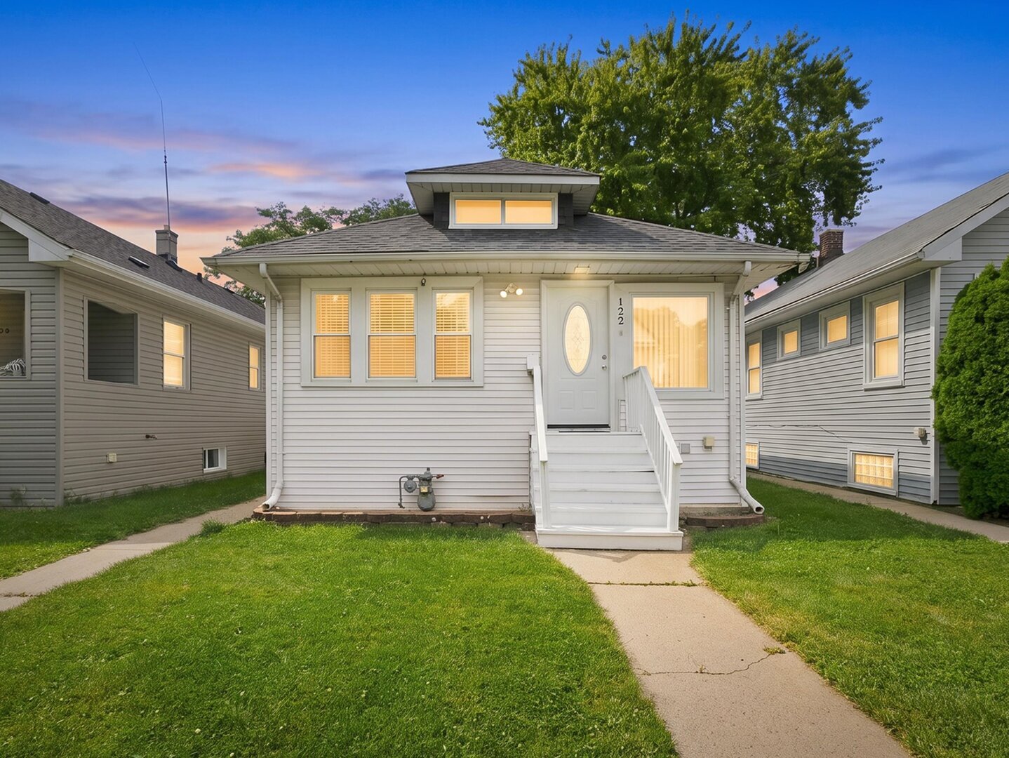 Light-gray house with white front steps, illuminated windows, and a broad lawn at dusk.
