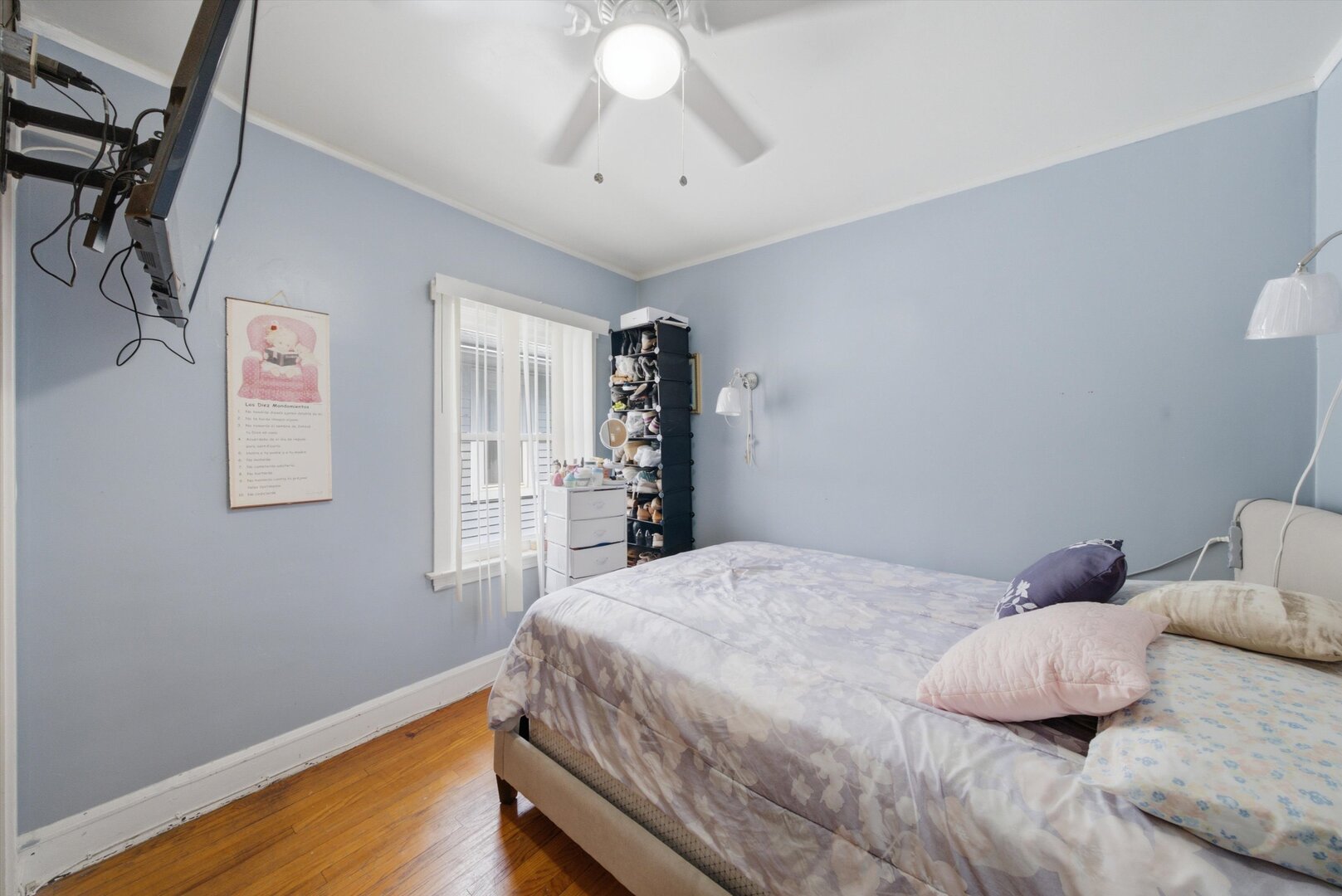 Bedroom with pale blue walls, a patterned bed, hardwood floors, and a window with vertical blinds.