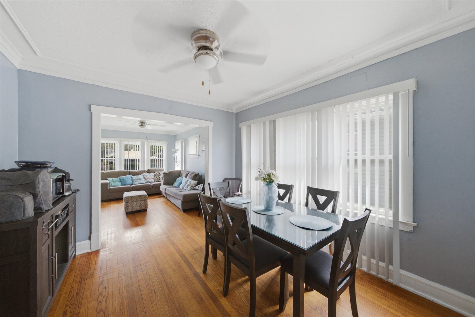 Dining room with a glass-top table and hardwood floors, opening to a living room with a gray sectional.