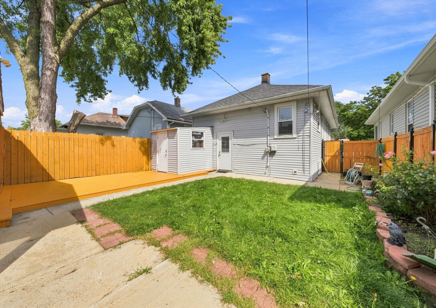 Gray-sided house behind a fenced backyard, with a wooden deck and concrete walkway.