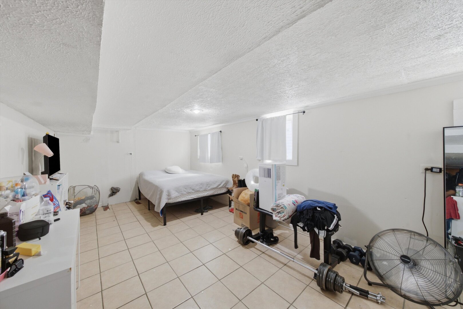 Bedroom with a bed, tiled floor, and exercise equipment including a barbell and large fan.