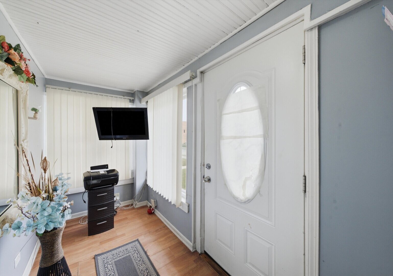 Entryway with a white oval-glass front door, light wood flooring, vertical blinds, and a wall-mounted TV.