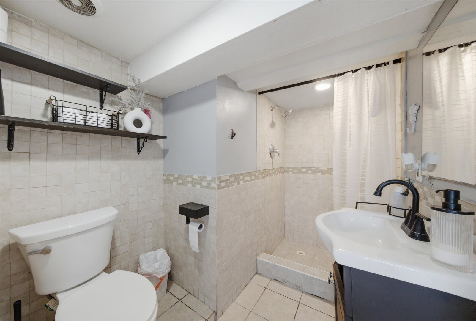 Tiled bathroom with a walk-in shower, white sink vanity, toilet, and dark wall shelves.