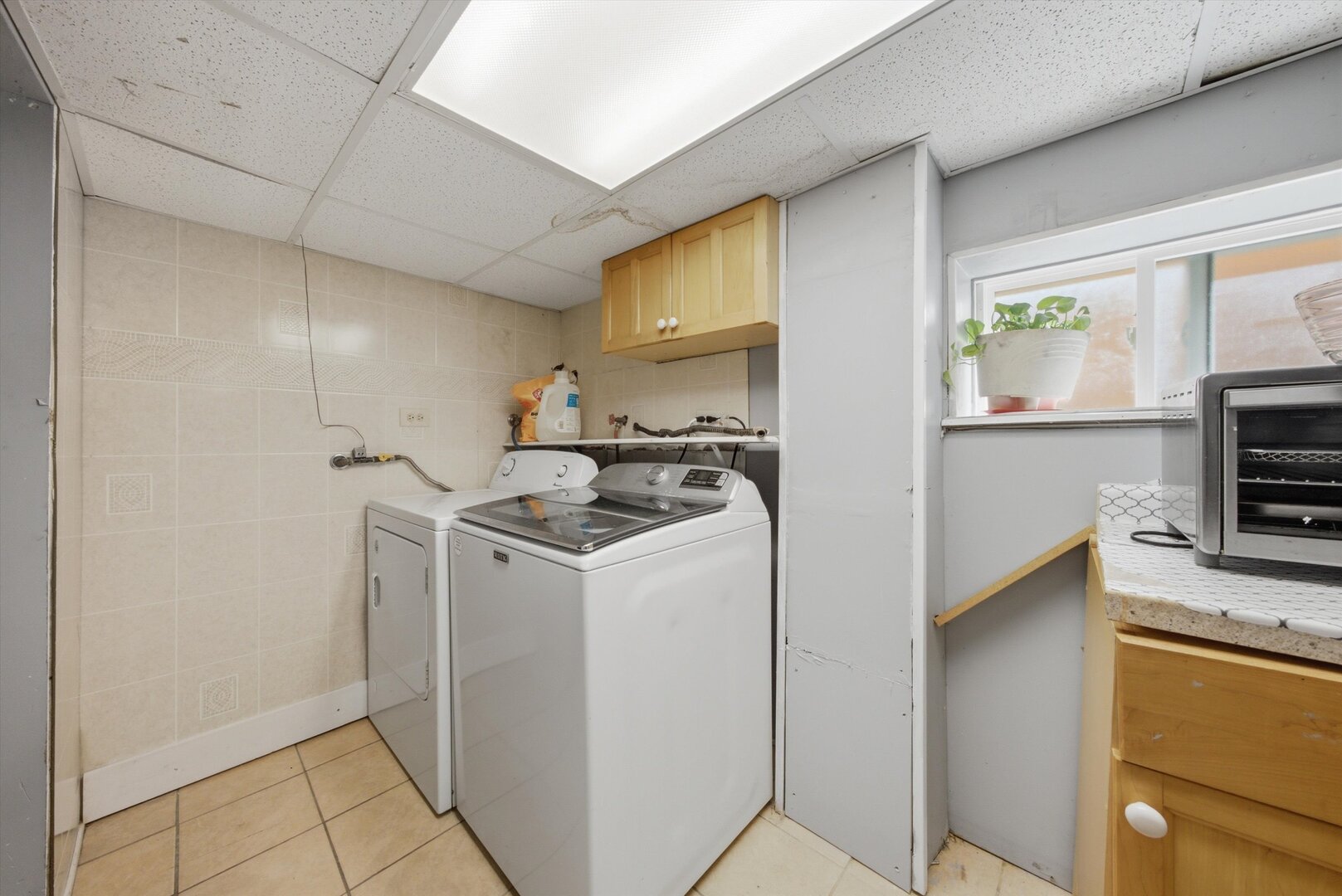 Laundry room with a white washer and dryer, tiled walls, and a light wood cabinet.