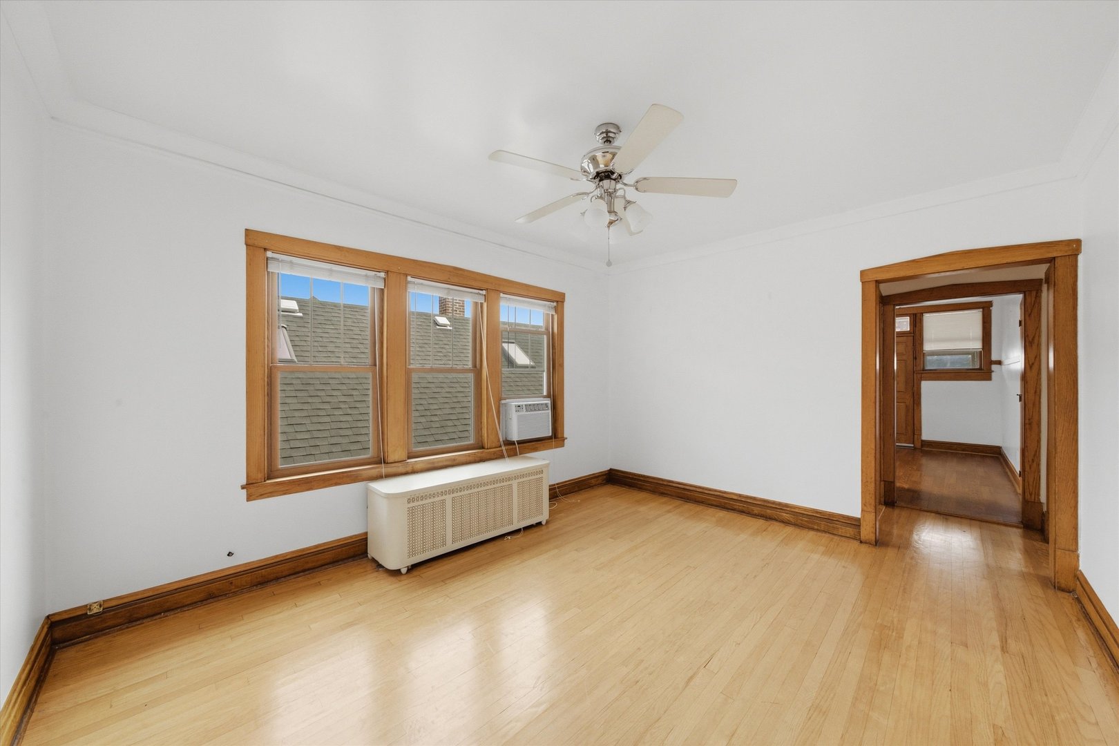 Vacant room with hardwood floors, wood-trimmed windows, ceiling fan, and radiator.