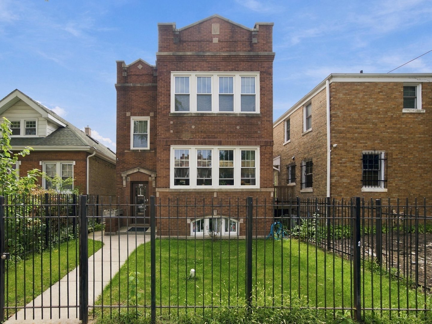 Red brick multi-story home with white-trimmed windows behind a black metal fence and small front lawn.