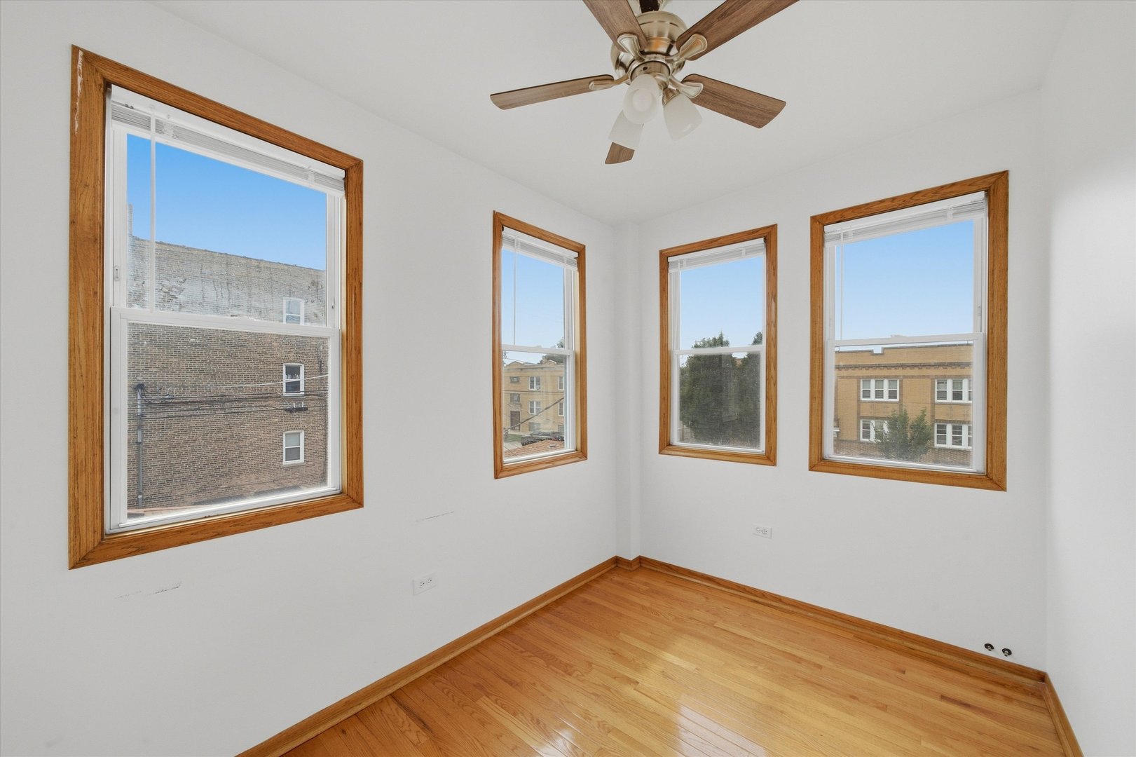 Empty room with hardwood floors, four wood-trimmed windows, and a ceiling fan