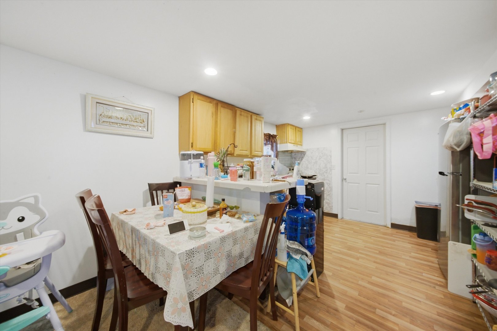 Dining area with a lace-covered table beside a kitchen with light wood cabinets and a white peninsula.