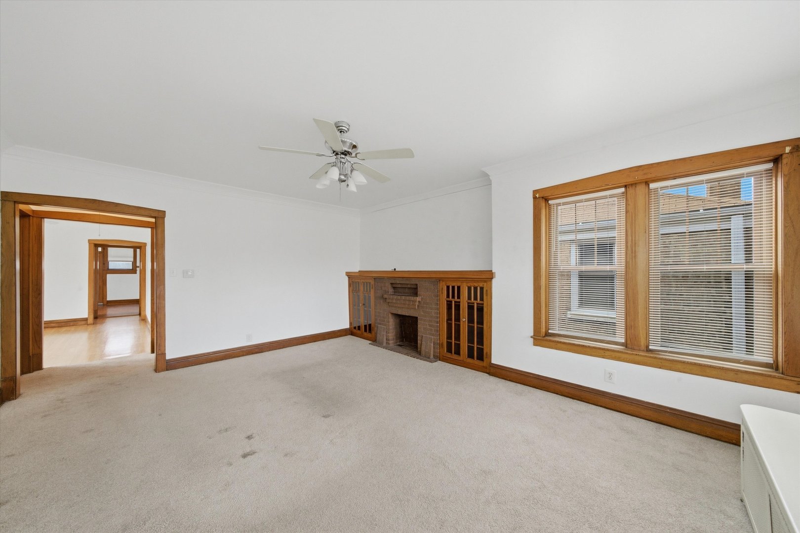 Empty living room with a brick fireplace, wood trim, large blinded windows, and a doorway to another room.