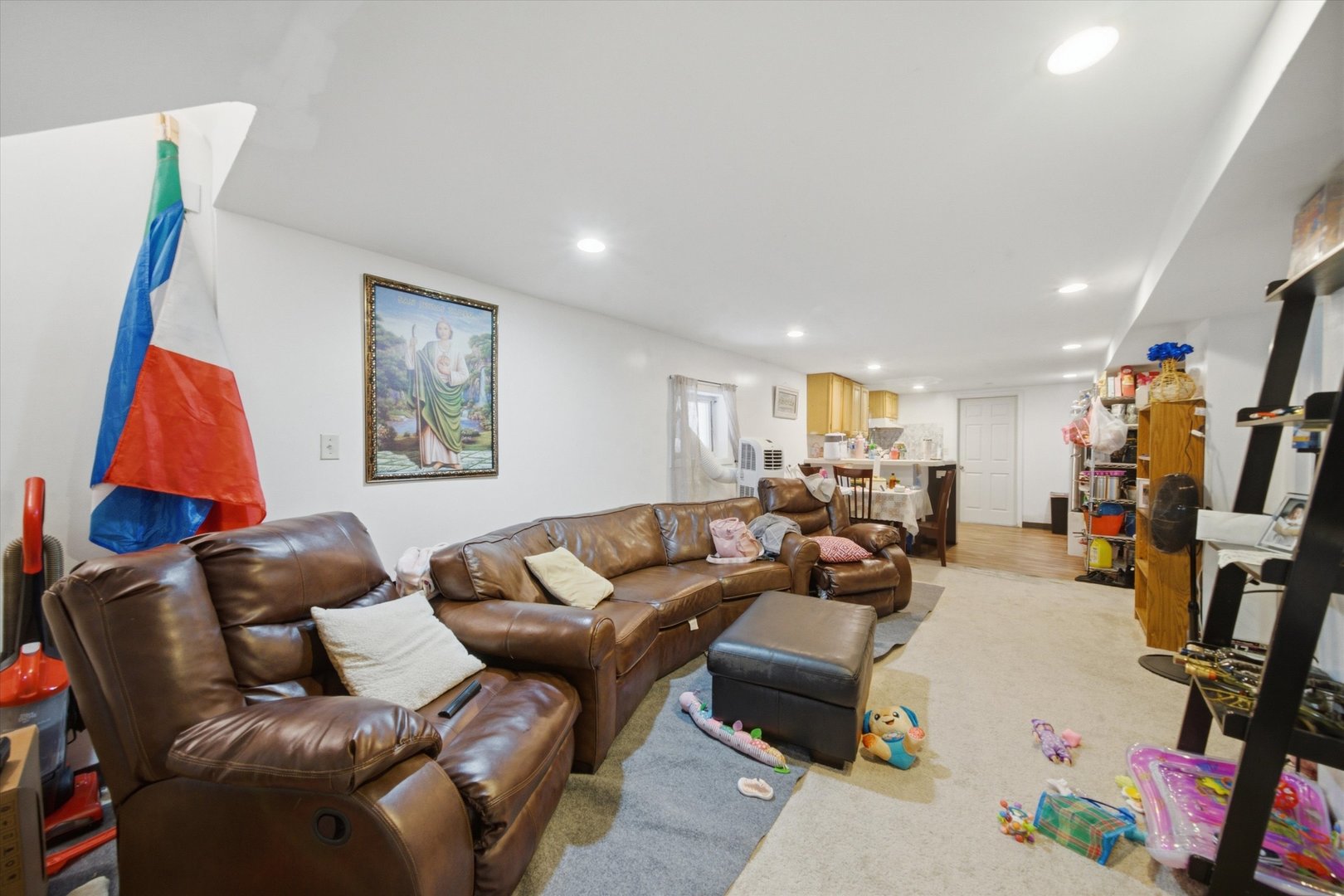 Furnished living room with brown leather seating, carpet, and an adjoining dining area and kitchen.