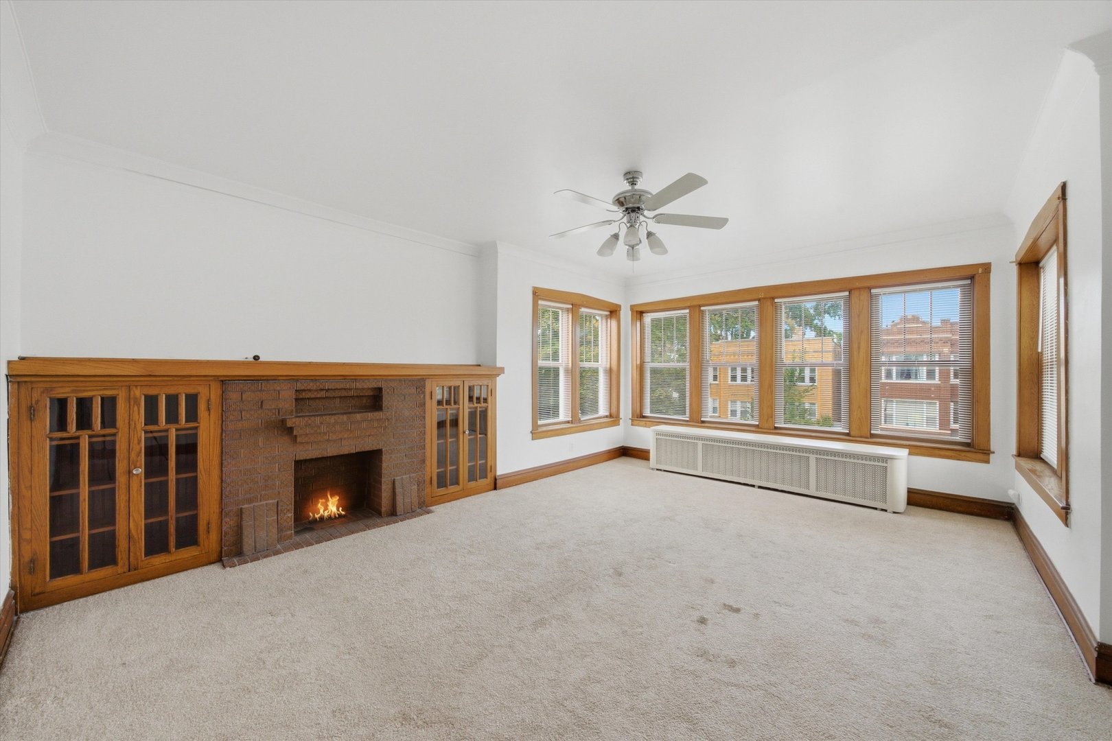 Empty living room with a brick fireplace, built-in wood cabinets, and broad windows.