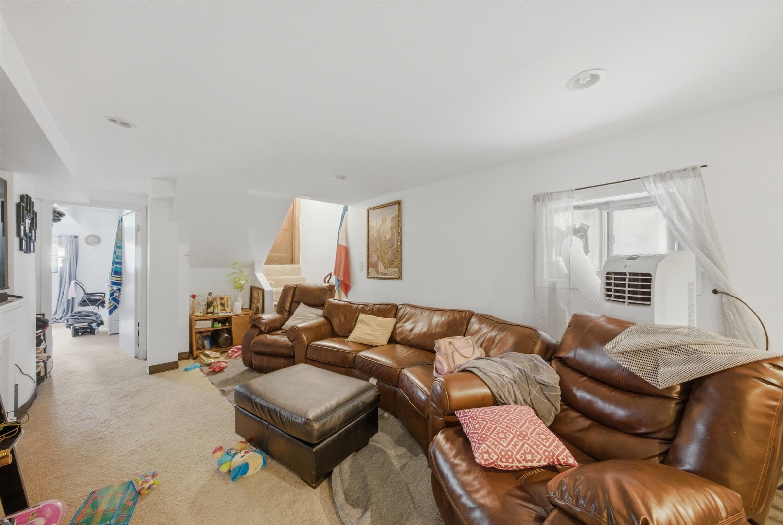 Living room with a large brown leather sectional, ottoman, staircase, and portable air conditioner.