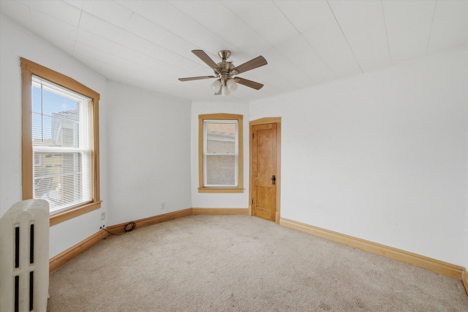 Vacant carpeted room with wood-trimmed windows, a wooden door, ceiling fan, and radiator.