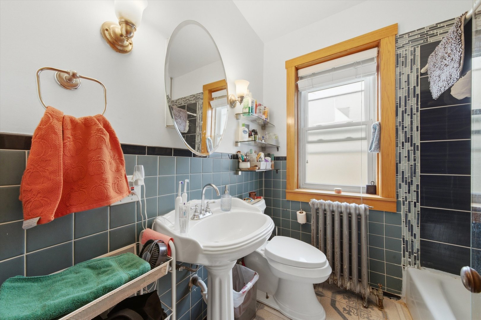 Bathroom with blue-gray tile, pedestal sink, wood-trimmed window, and dark mosaic accent wall.