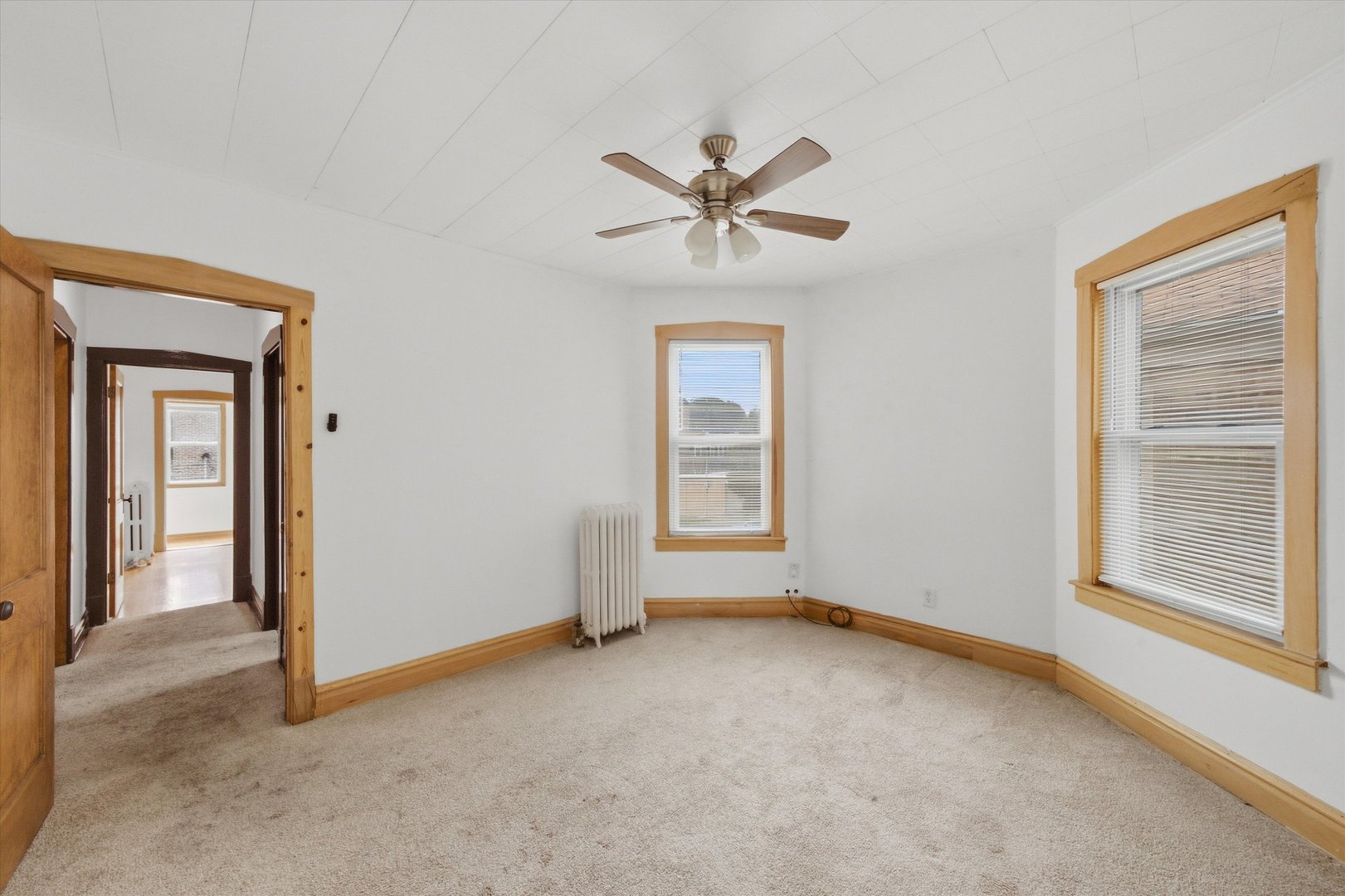 Empty carpeted bedroom with wood-trimmed windows, a ceiling fan, and a radiator.