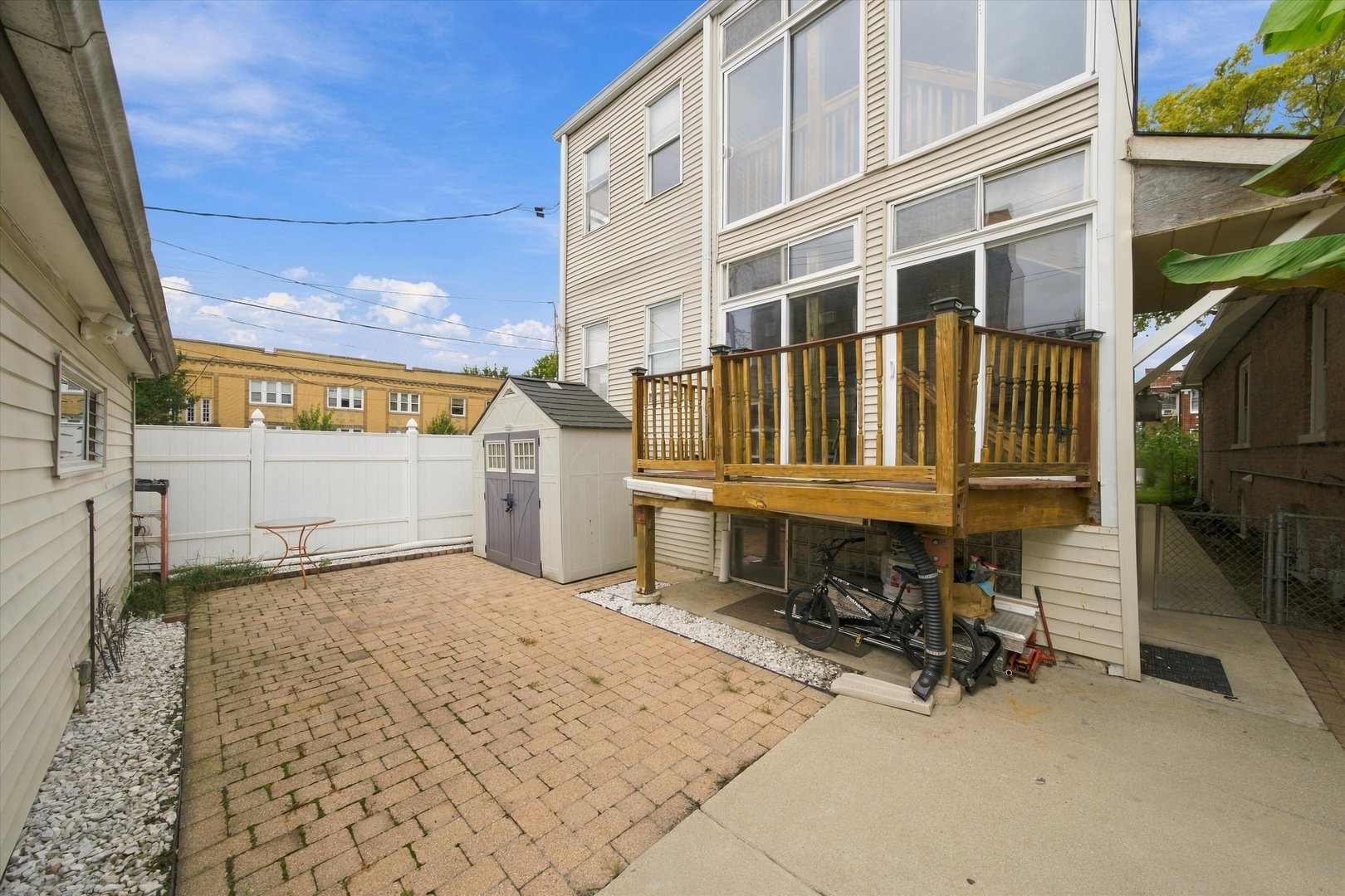 Backyard patio with brick pavers, a raised wooden deck, white fence, and storage shed.