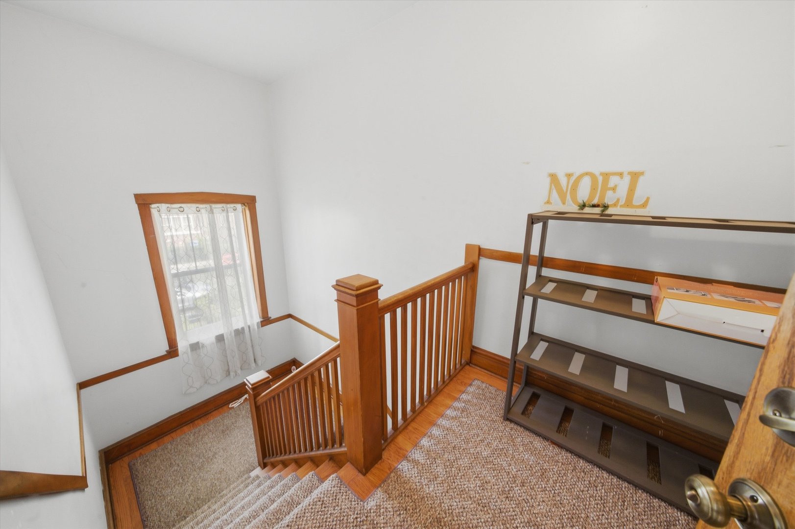 Carpeted staircase and landing with wood railings, a curtained window, and a shelf displaying “NOEL” letters.
