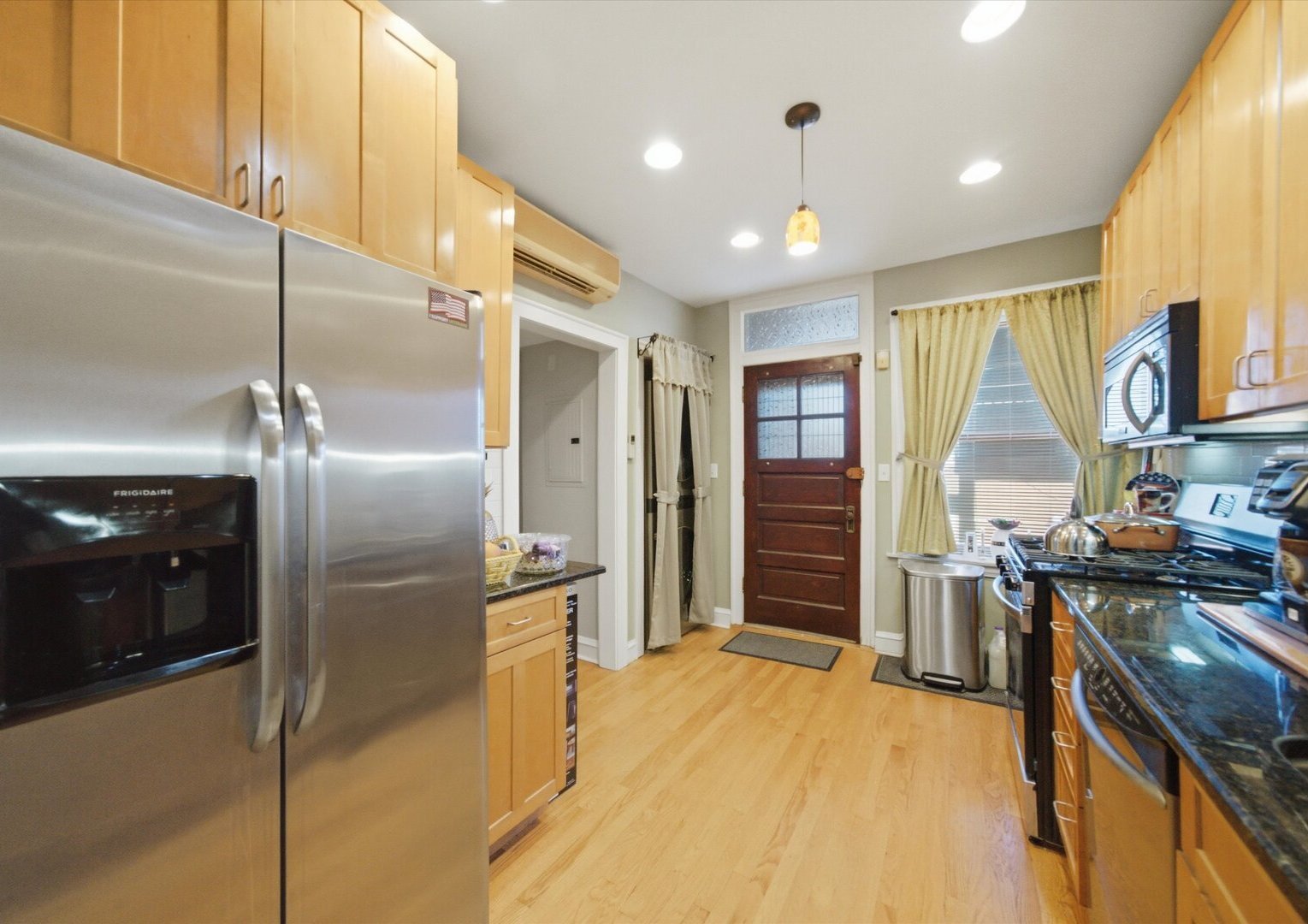 Kitchen with light wood cabinets, stainless steel appliances, and a dark wood entry door.