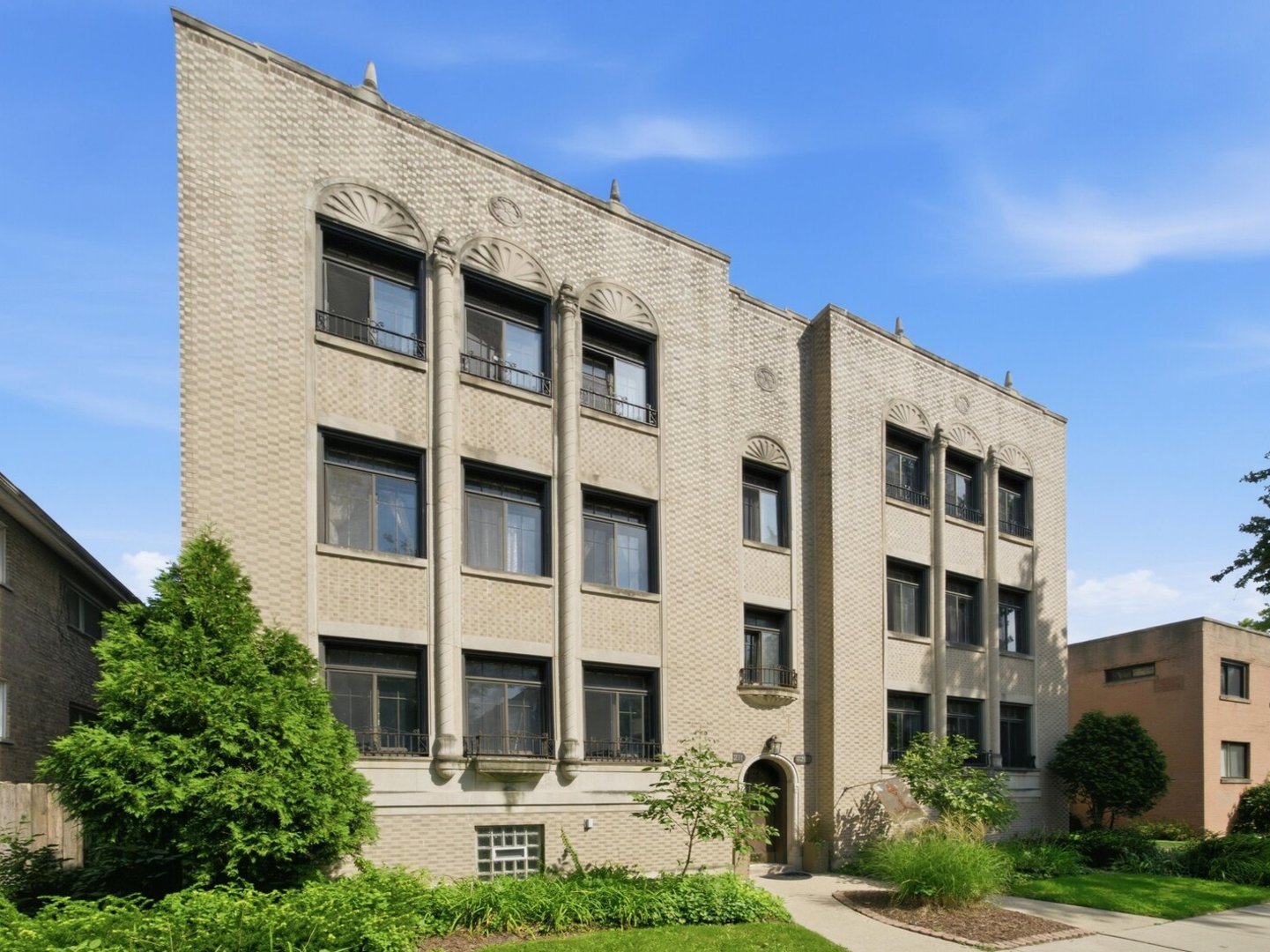 Beige brick apartment building with decorative arched windows and landscaped grounds.