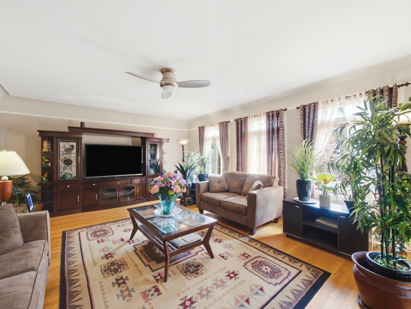 Living room with hardwood floors, a patterned rug, dark wood entertainment center, and large curtained windows.