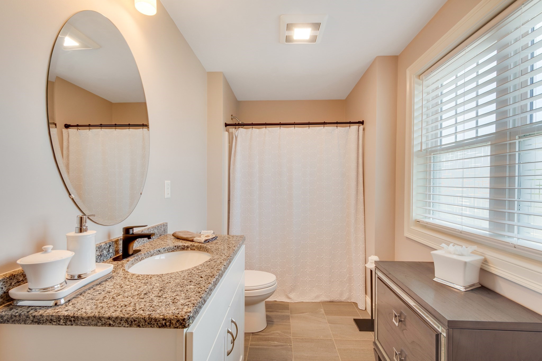 Bathroom with a granite vanity, oval mirror, and window beside the curtained tub.
