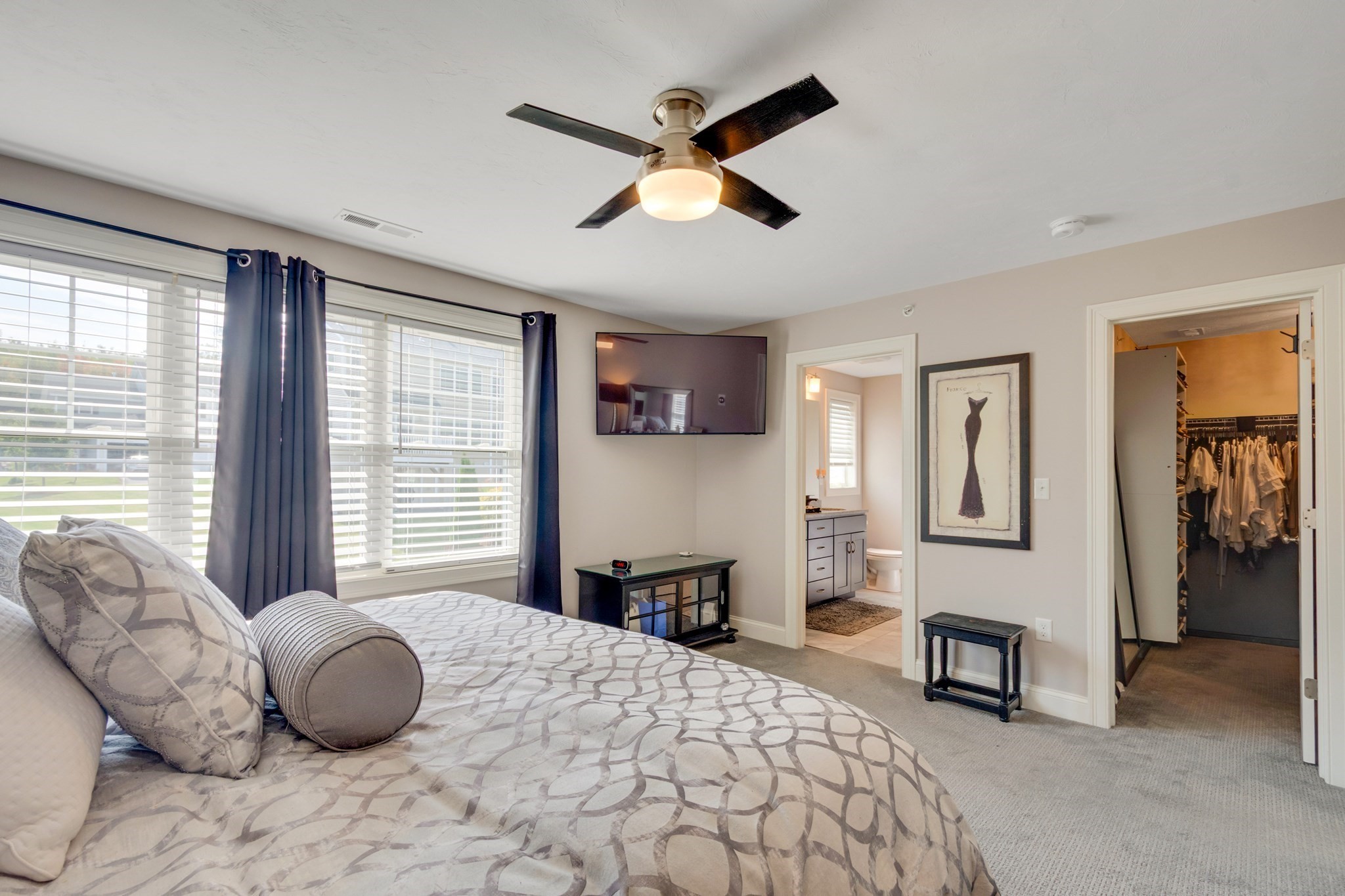 Furnished bedroom with gray patterned bedding, navy curtains, and open doors to a bathroom and walk-in closet.