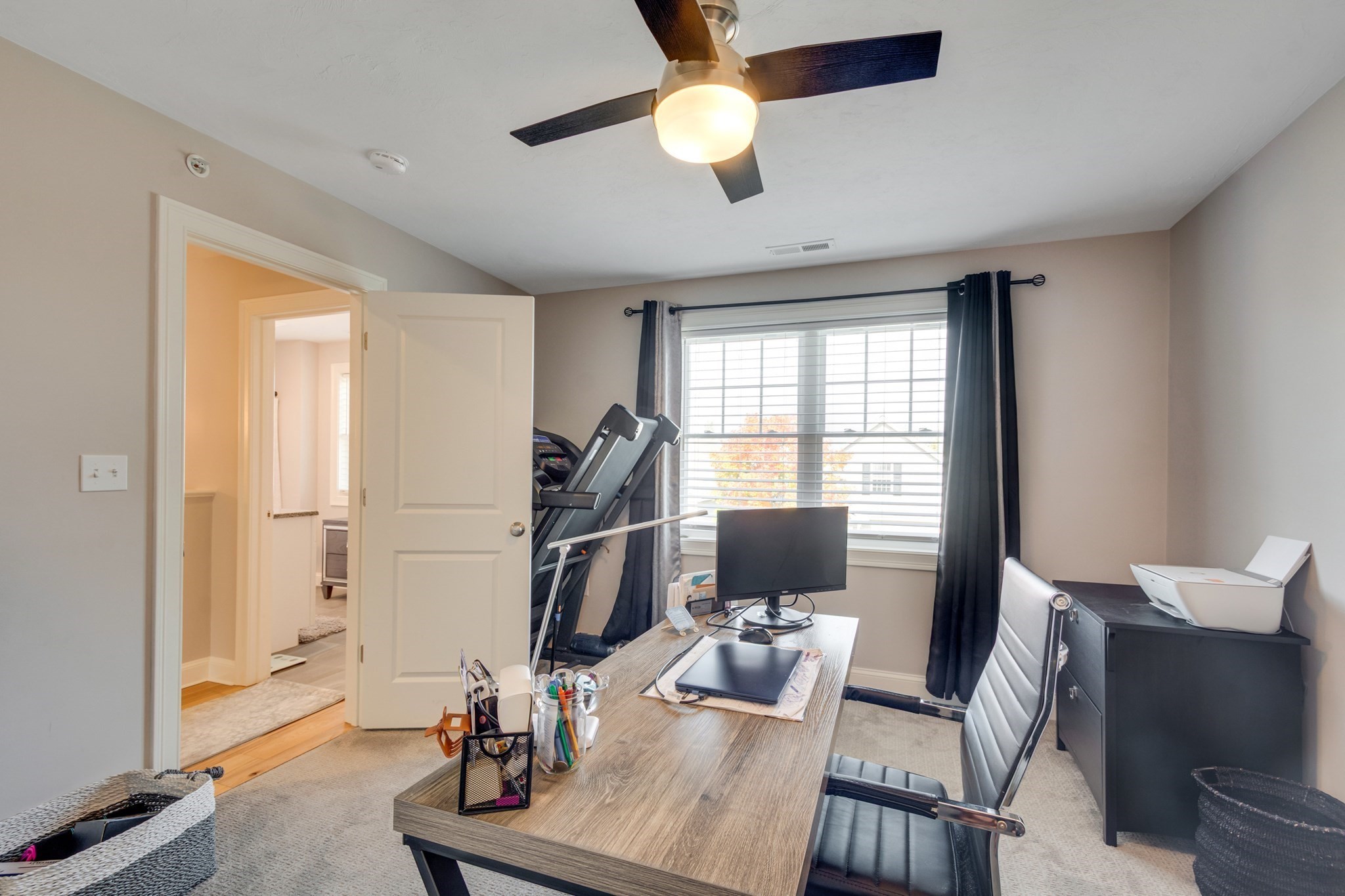 Home office with a wood desk, treadmill beside a curtained window, and ceiling fan.