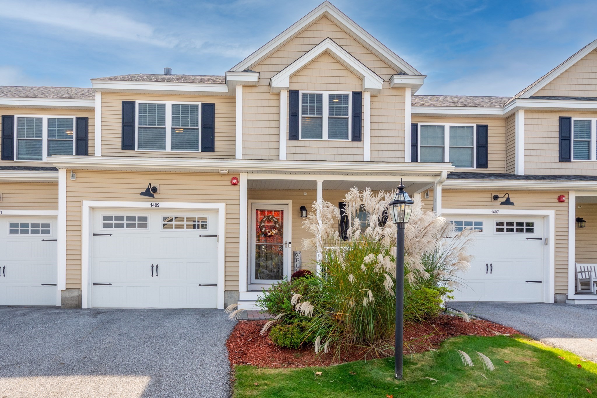 Beige two-story townhouse with dark shutters, a white garage door, and ornamental grasses by the entry.