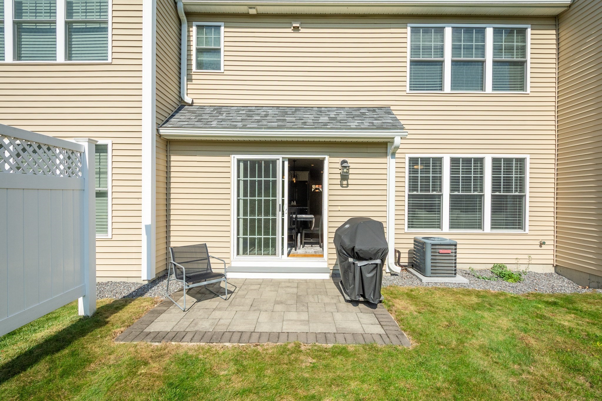 Home’s rear exterior with a paved patio, sliding glass door, lawn, and white fence.