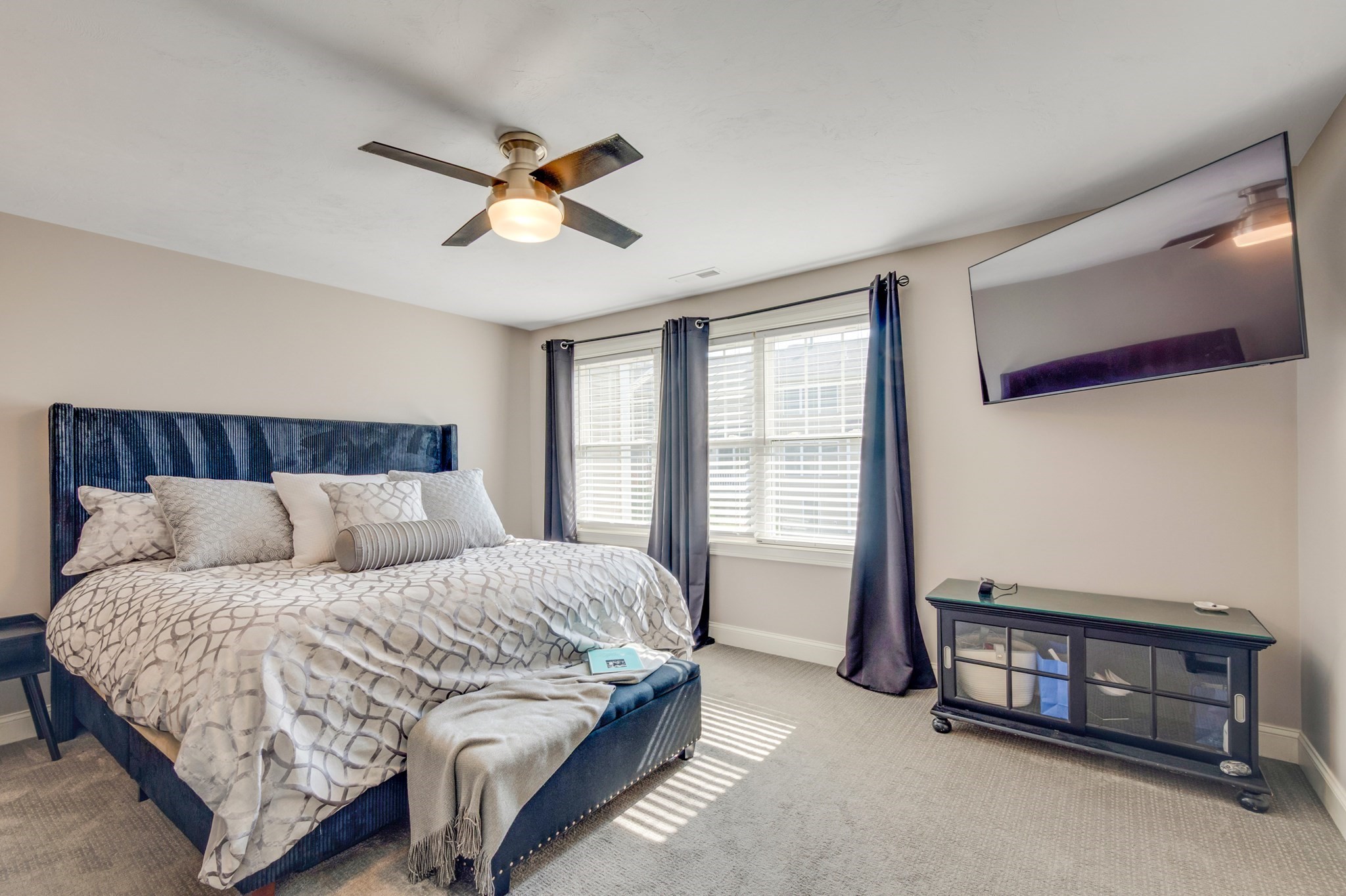 Bedroom with a navy upholstered bed, patterned bedding, sunny window, and wall-mounted television.