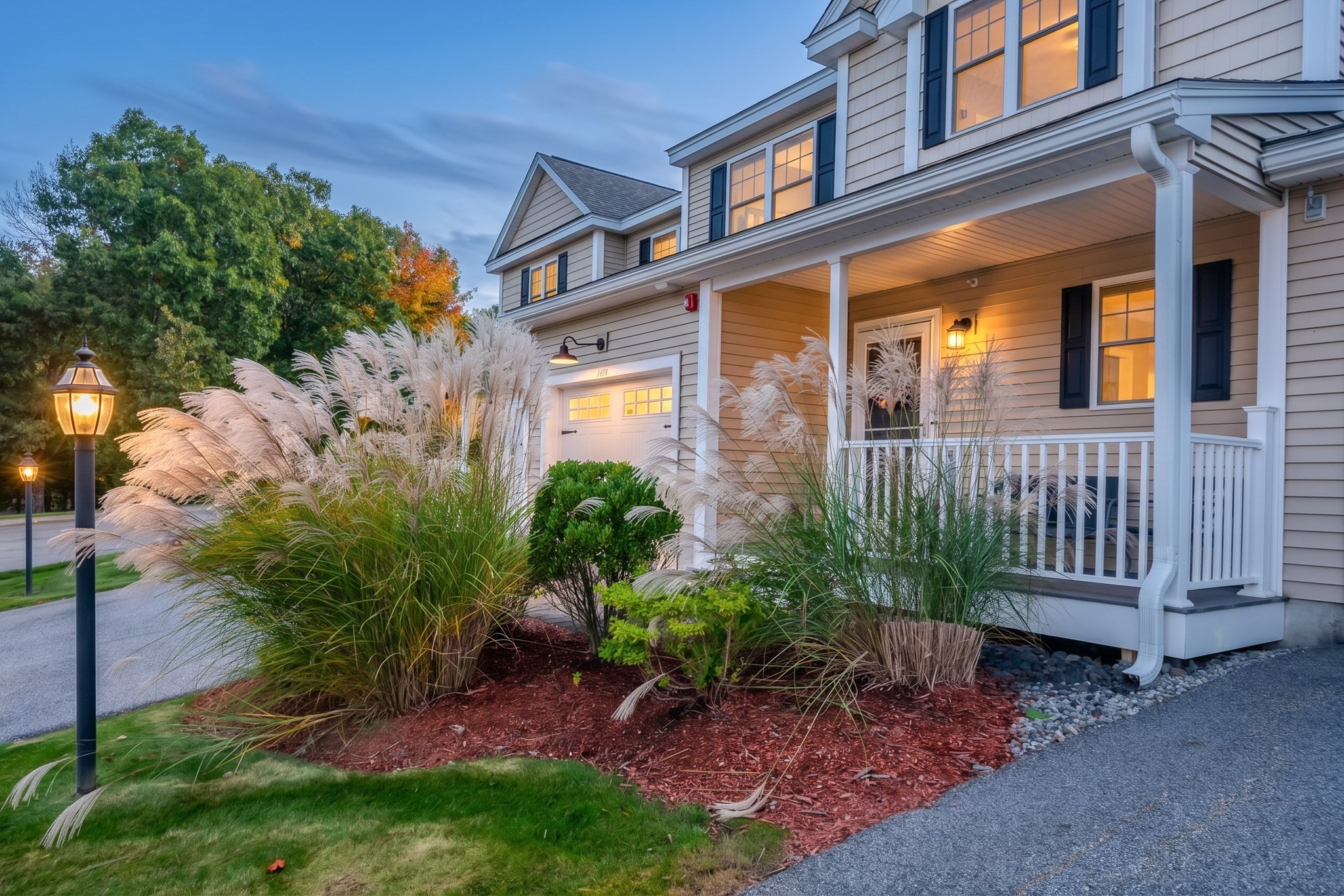 Two-story gray house with a covered porch, glowing windows, and ornamental grasses in the front garden.