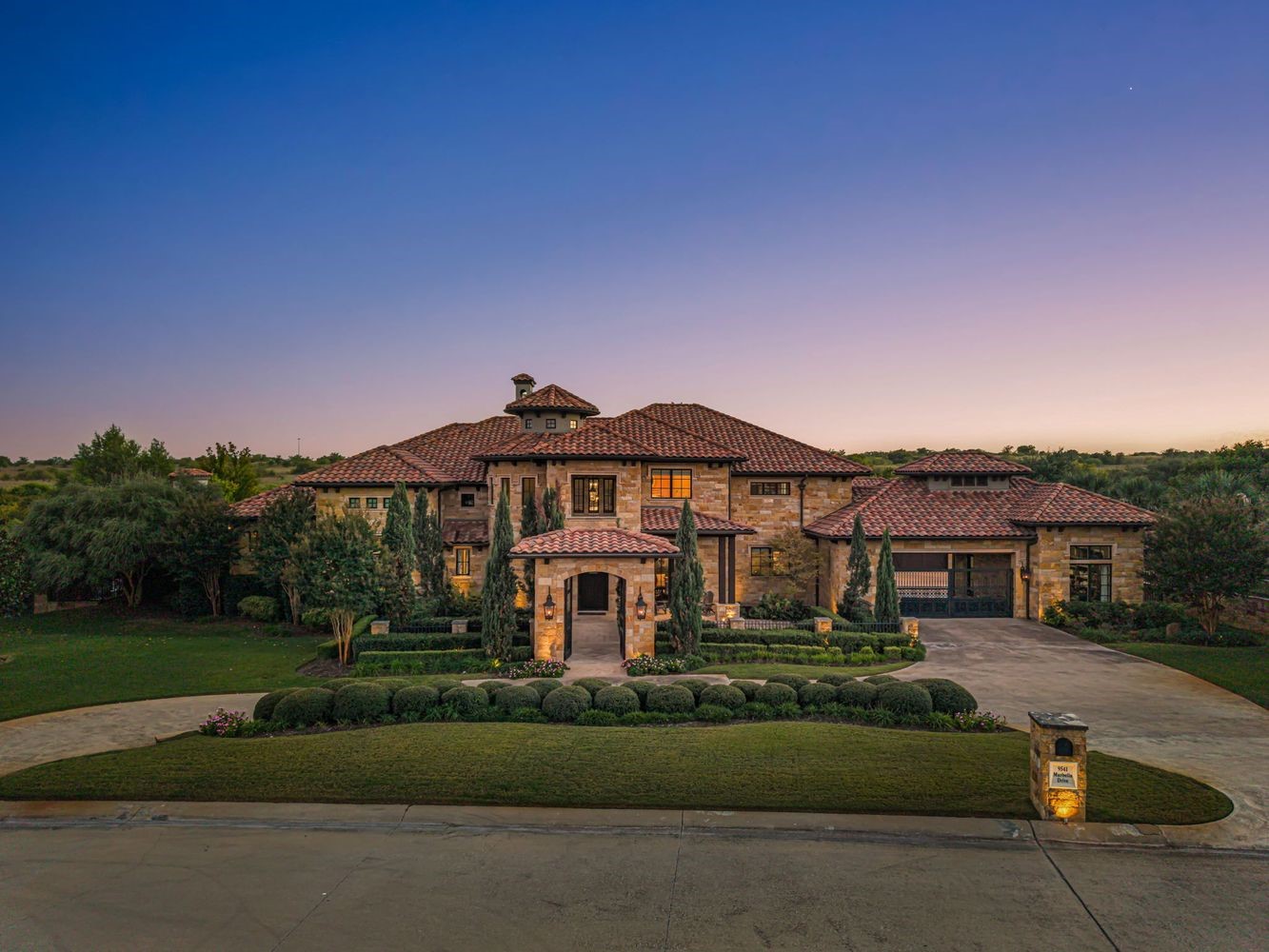 Stone house with a red tile roof, landscaped lawn, and curved driveway at dusk.