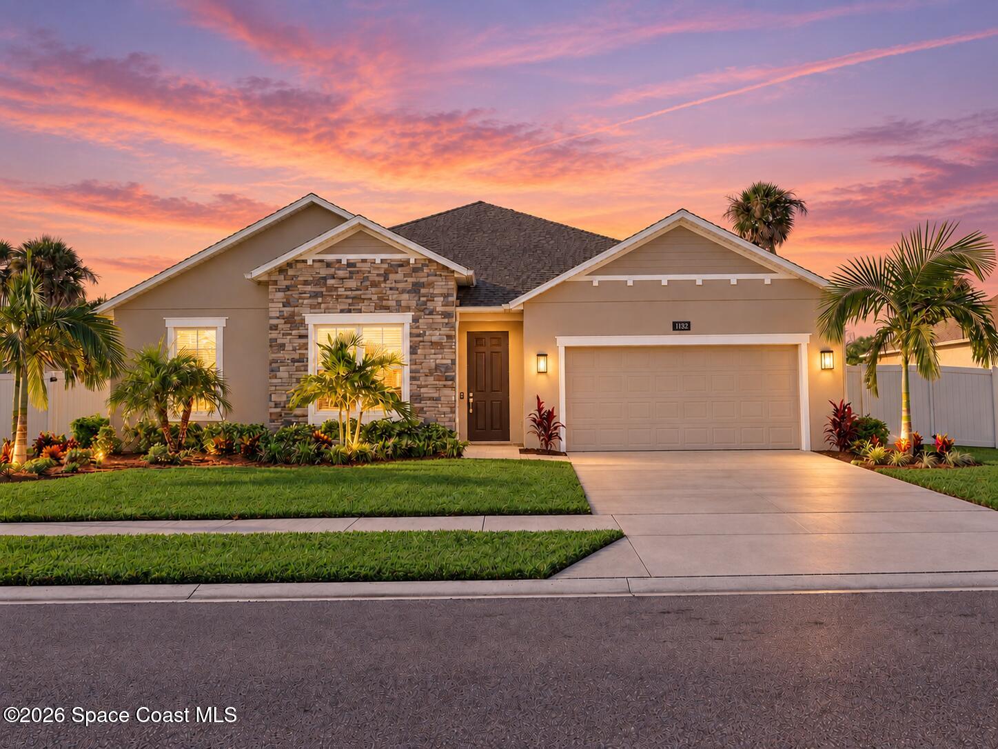 Home with stone facade, two-car garage, and tropical landscaping beneath a colorful sunset sky.