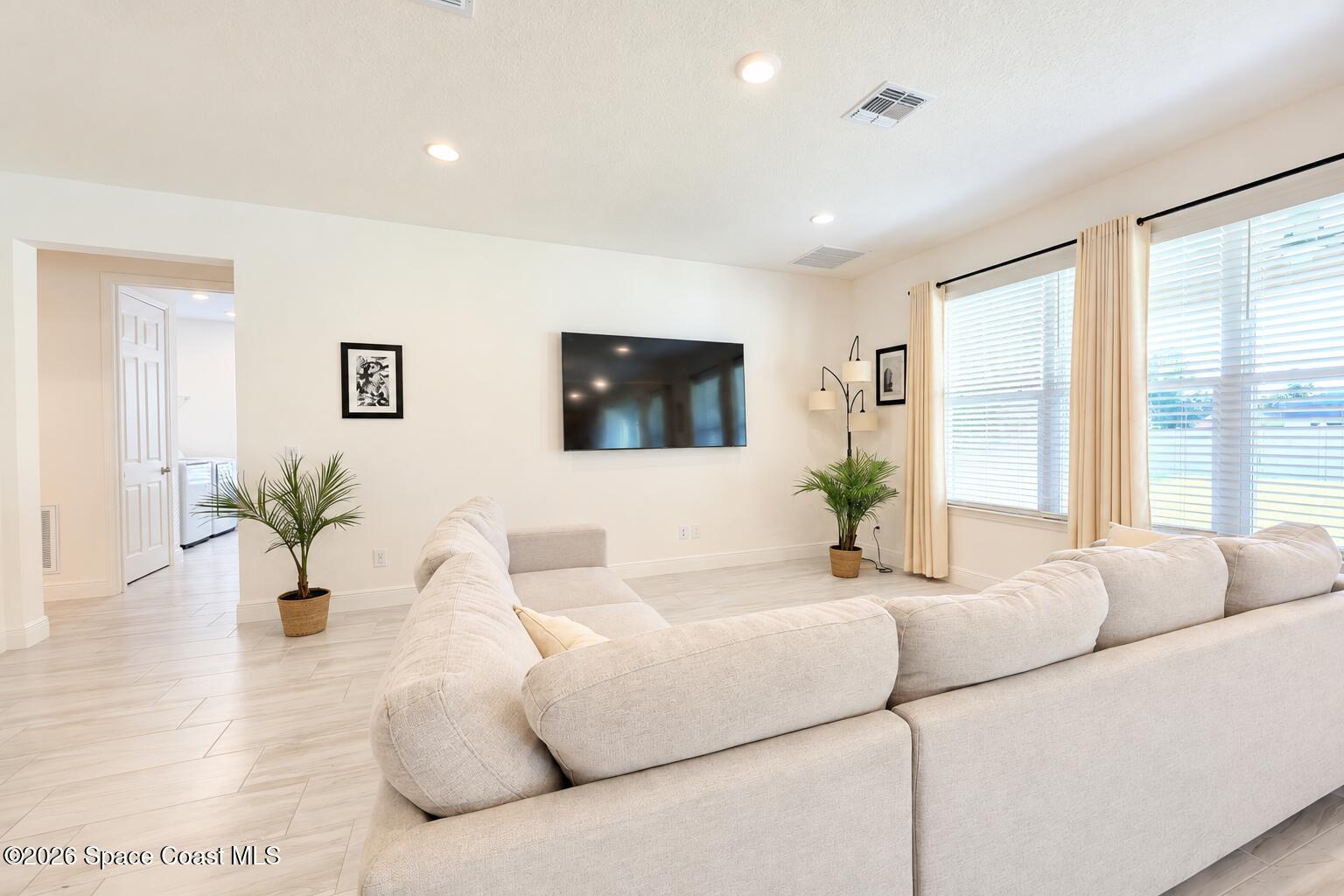 Bright living room with a cream sectional, wall-mounted TV, tall curtained windows, and pale tile flooring.