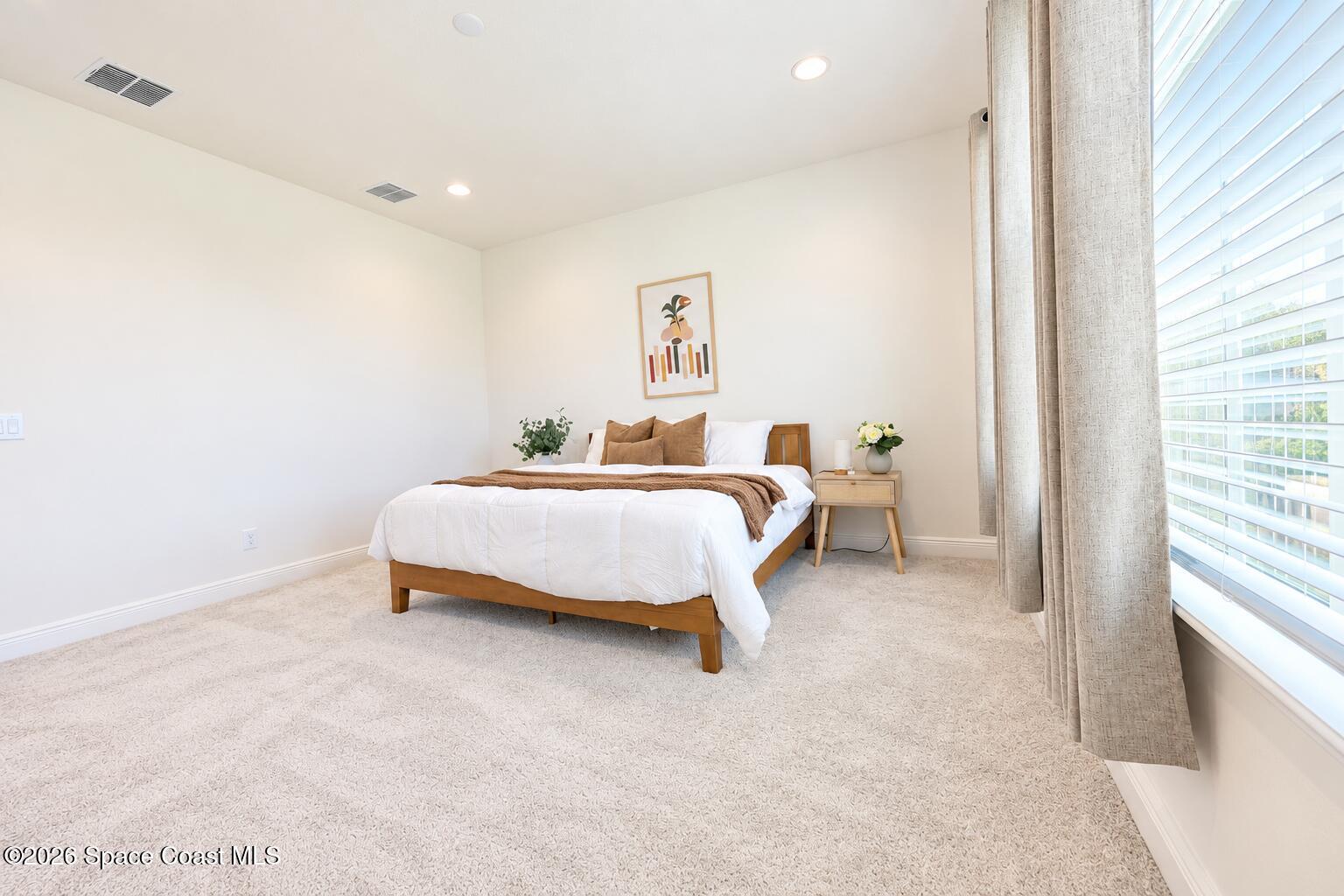 Staged bedroom with a wooden bed, white bedding, beige carpet, and a large curtained window.