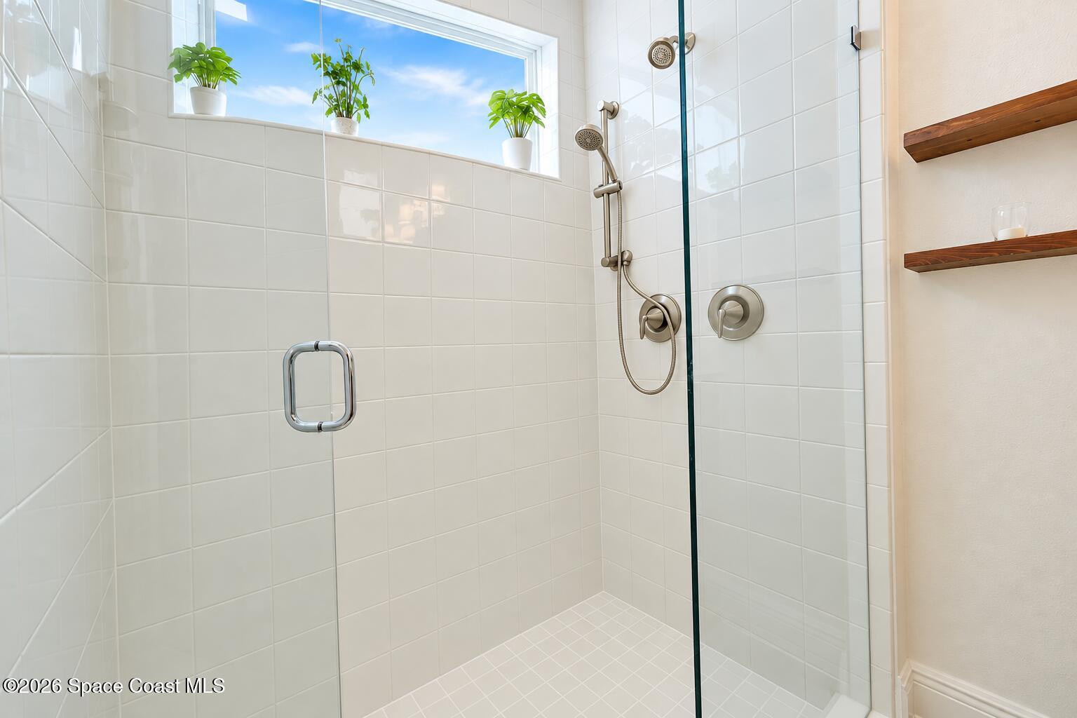 White-tiled walk-in shower with glass enclosure, brushed-metal fixtures, and a high window with potted plants.