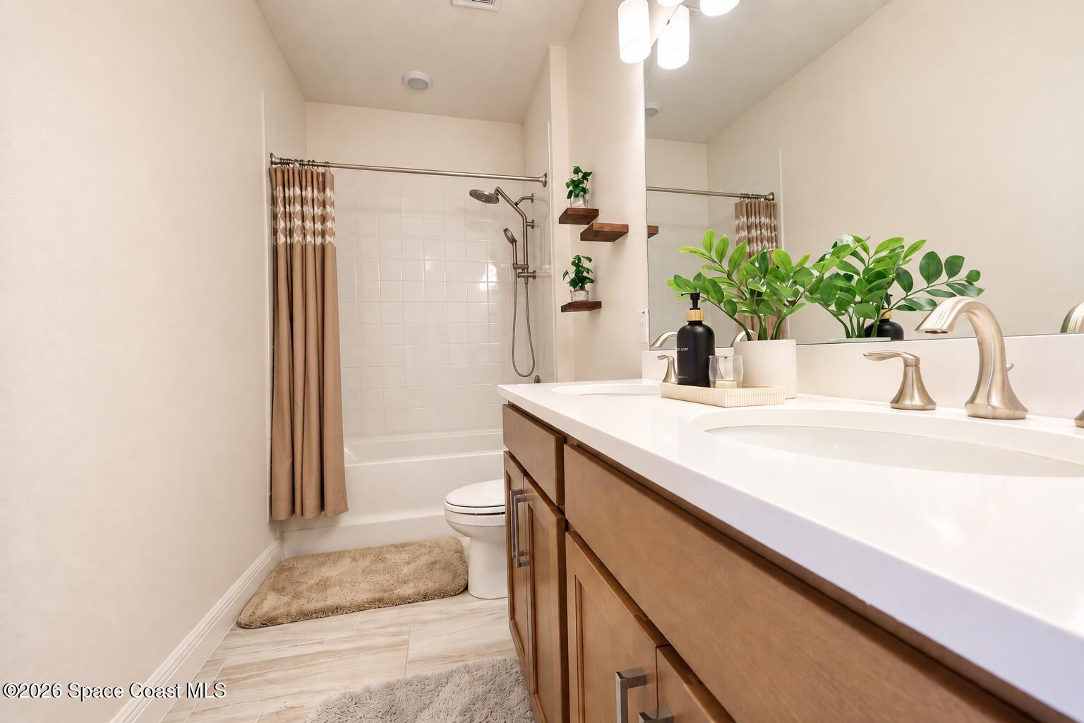 Bathroom with a double-sink vanity and wood cabinets beside a tub-shower with a patterned curtain.