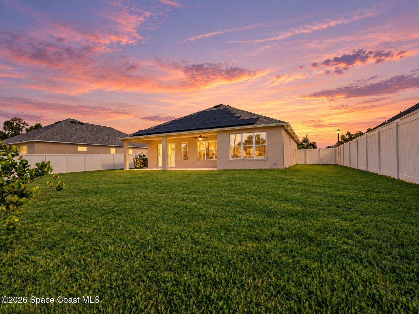 Rear of a single-story home with rooftop solar panels, fenced lawn, and vivid sunset sky.