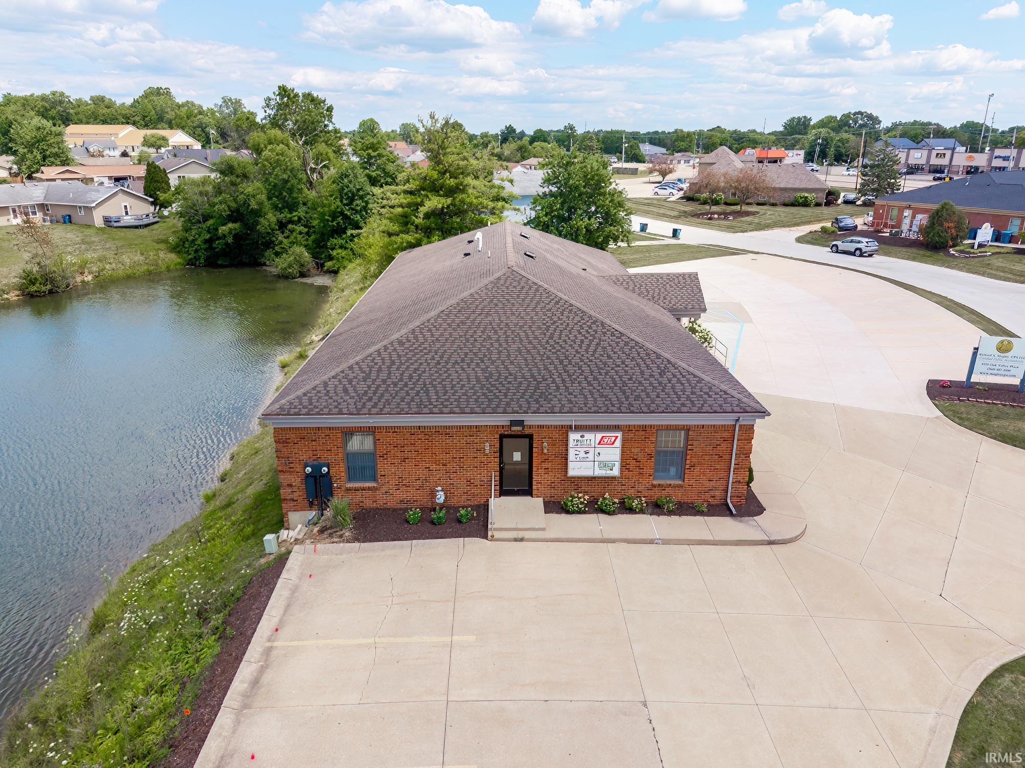 Aerial view of a red-brick commercial building beside a pond, with a wide concrete parking area.