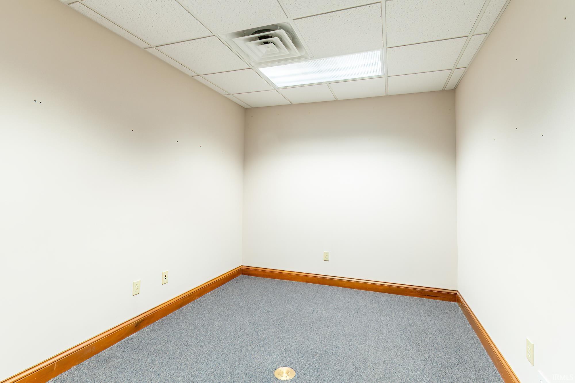 Empty carpeted room with wood baseboards, a drop ceiling, and a floor outlet.