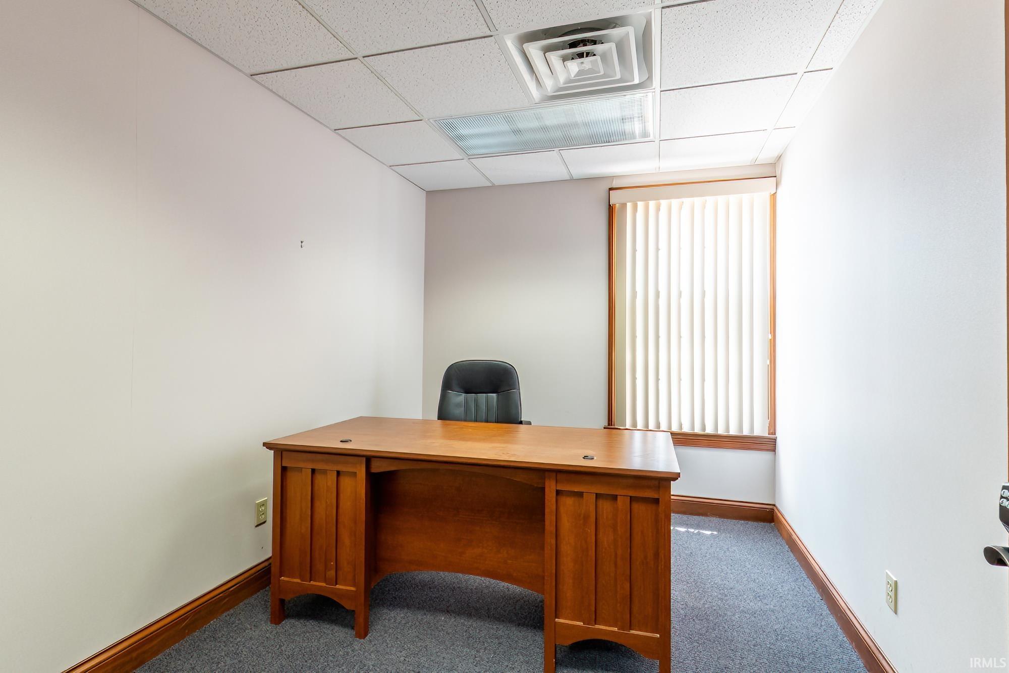 Small office with a wooden desk, black chair, vertical blinds, and gray carpet.