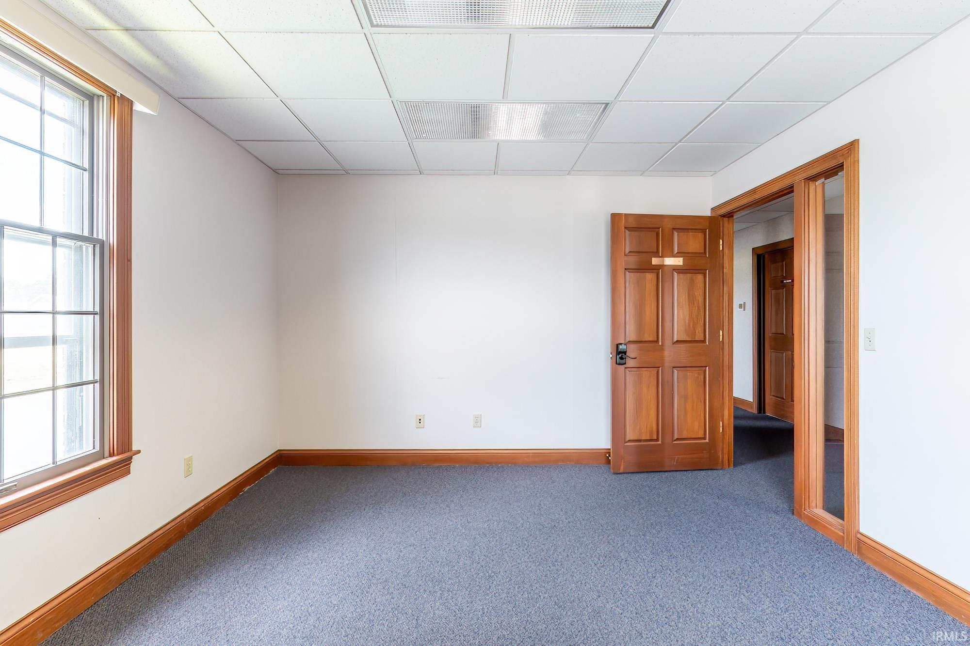 Empty office with blue-gray carpet, wood trim, a large window, and an open door to the hallway.