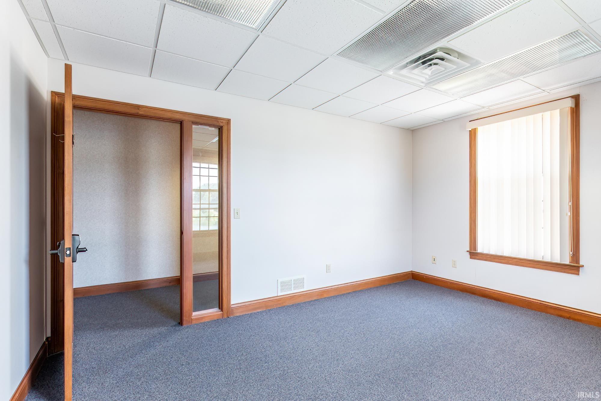 Vacant office with blue-gray carpet, wood trim, vertical blinds, and an open glass-sided doorway.