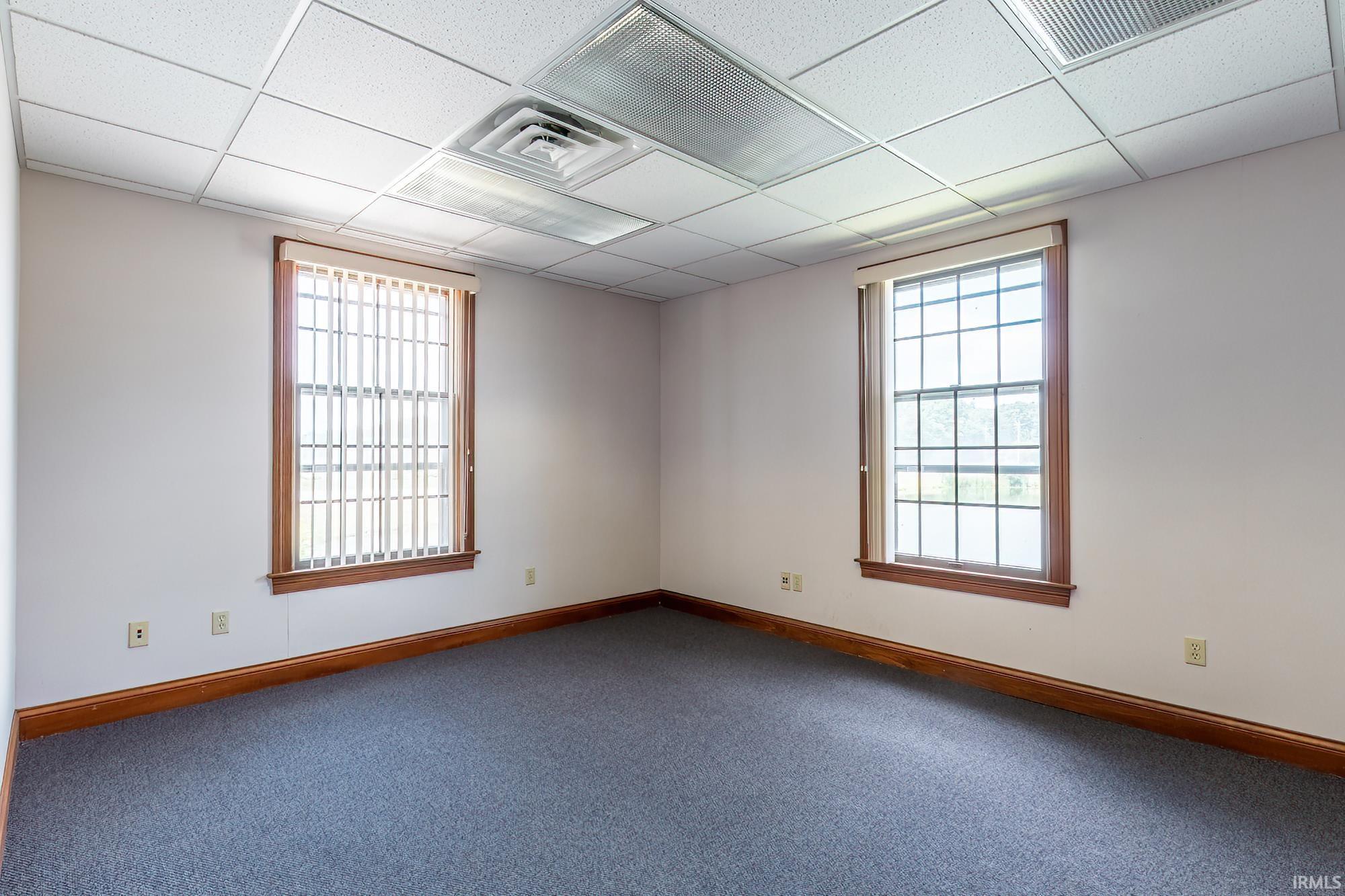 Vacant office with gray carpet, wood-trimmed windows, and a suspended ceiling.
