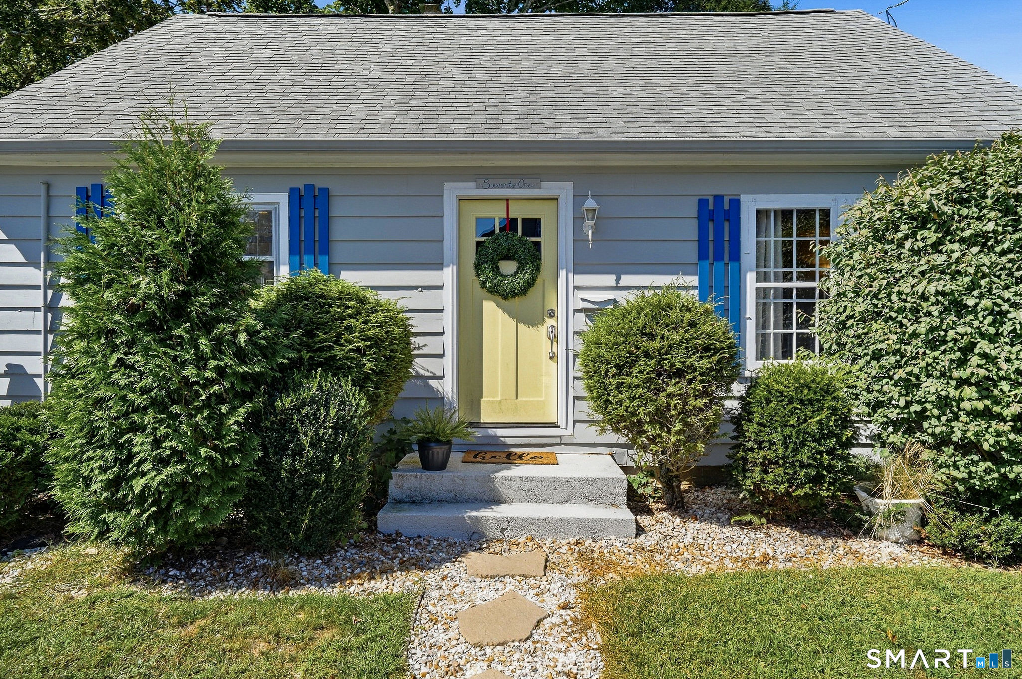 Gray house with blue shutters and a yellow, wreath-adorned front door, surrounded by shrubs.