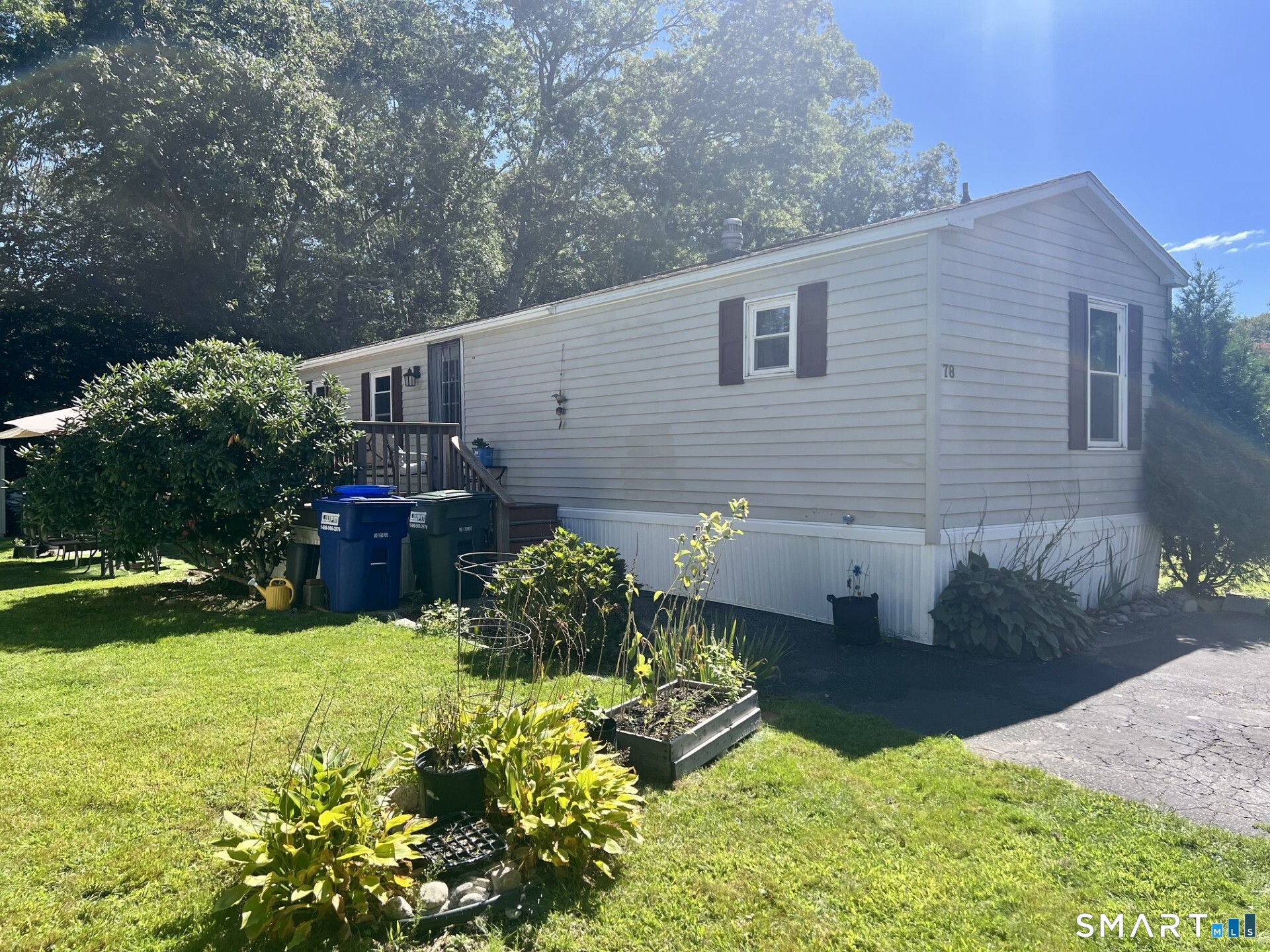 White-sided single-story home with a small deck, garden beds, and mature trees beside the lawn.