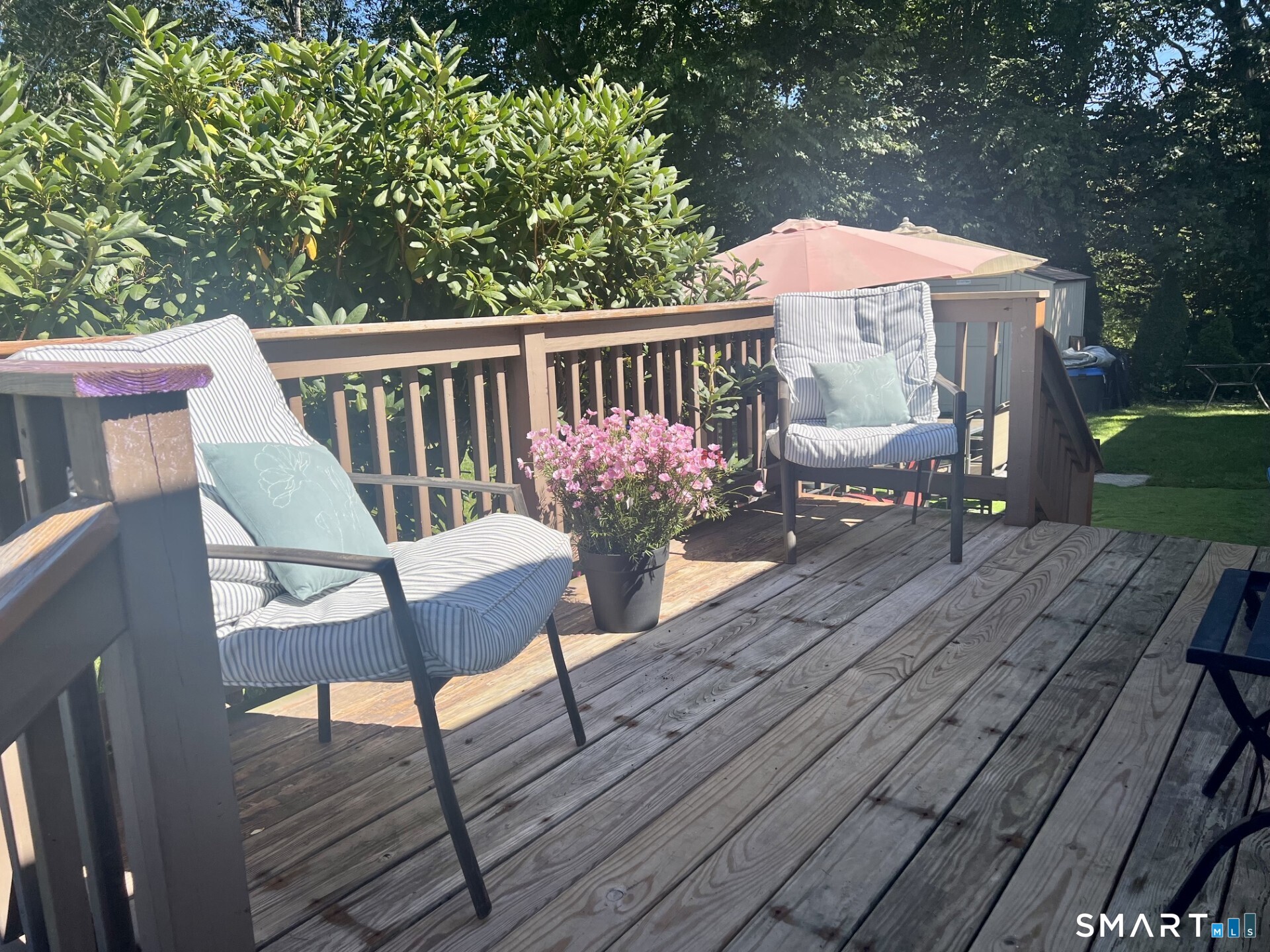 Wooden backyard deck with cushioned chairs, a potted pink flowering plant, and a patio umbrella.