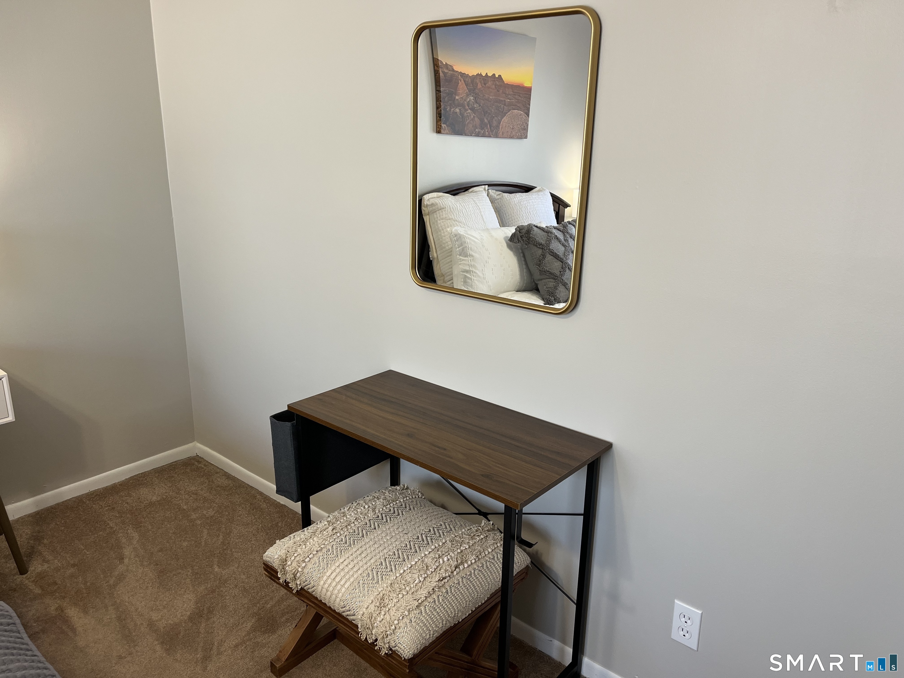 Bedroom corner with a wood desk, cushioned stool, and mirror reflecting the bed.