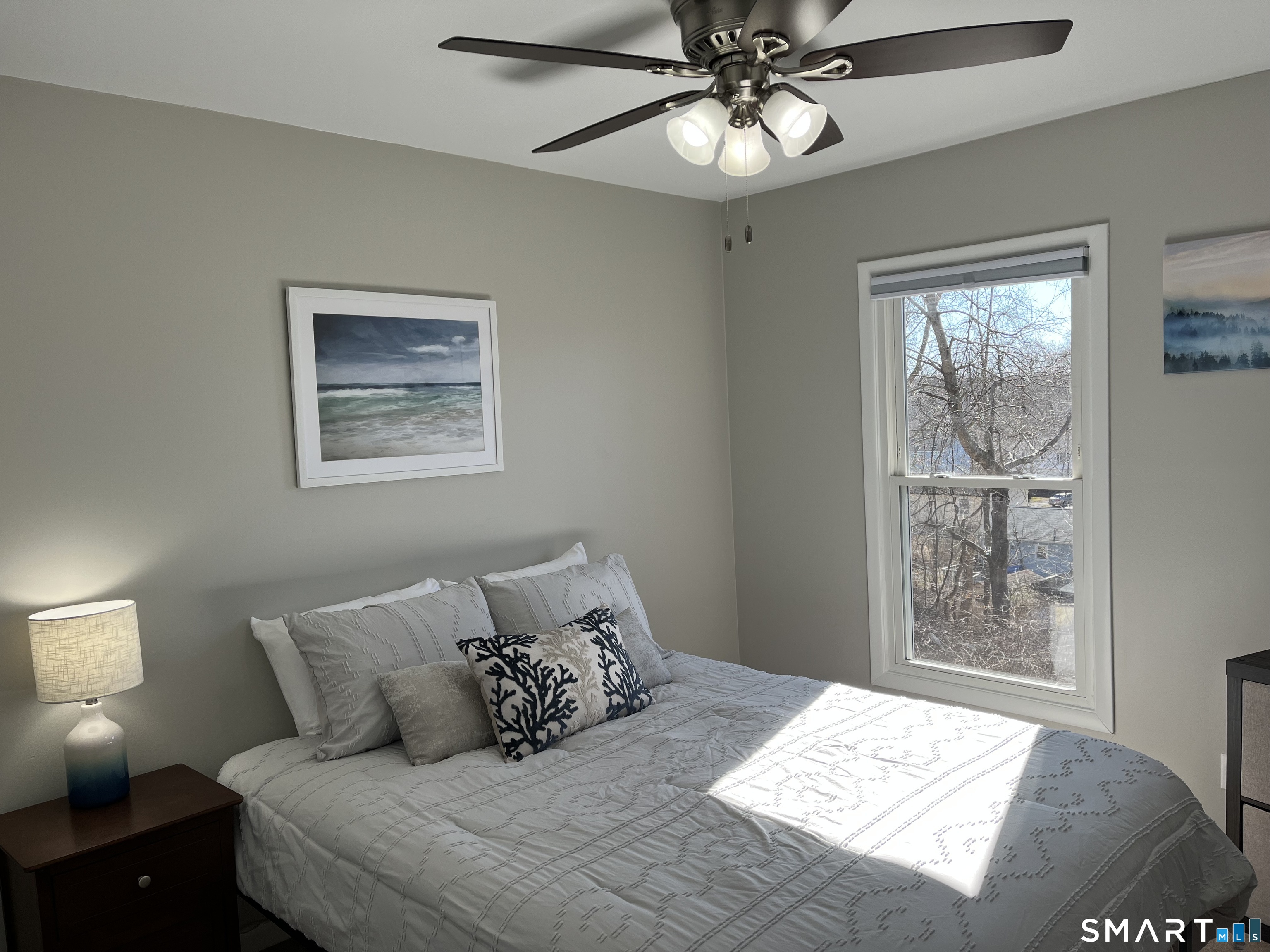 Furnished bedroom with a gray bed, ceiling fan, and sunny window overlooking leafless trees.